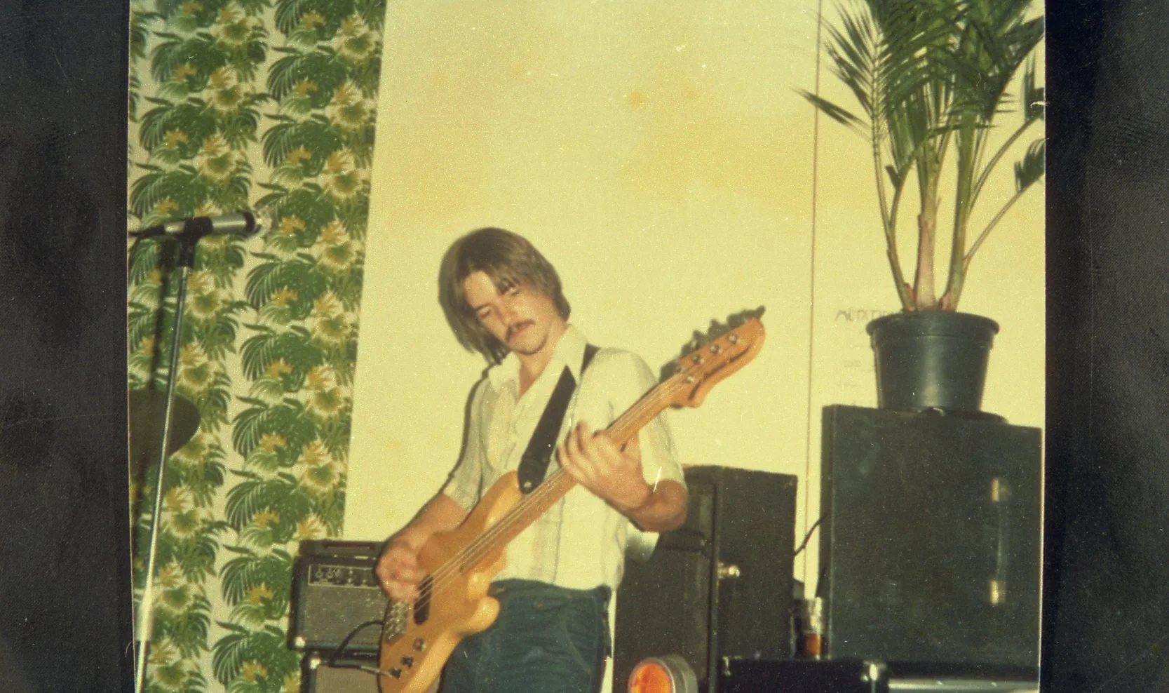A young man with shoulder-length brown hair performing with an acoustic guitar on a stage. He is dressed in a white short-sleeve button-up shirt with black accents, standing in front of a green floral curtain and a yellow wall with a potted plant on 