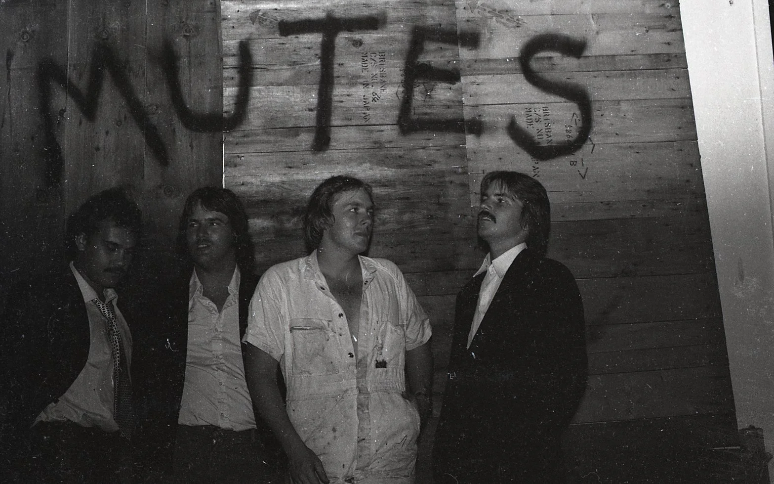 Black and white photo of four men standing in front of a wooden wall with the word 'HITS' spray-painted in large letters above them.