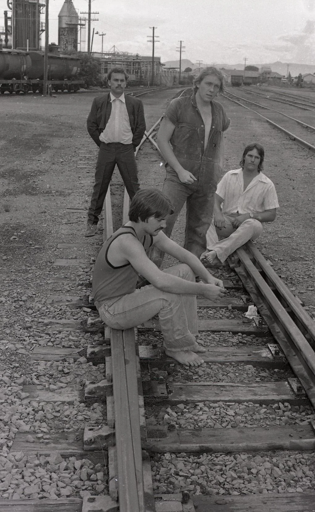 Four young men on a railway track with industrial buildings and water towers in the background. Two of them are sitting on the track, one is standing with his hands in pockets, and one is leaning against the tracks.