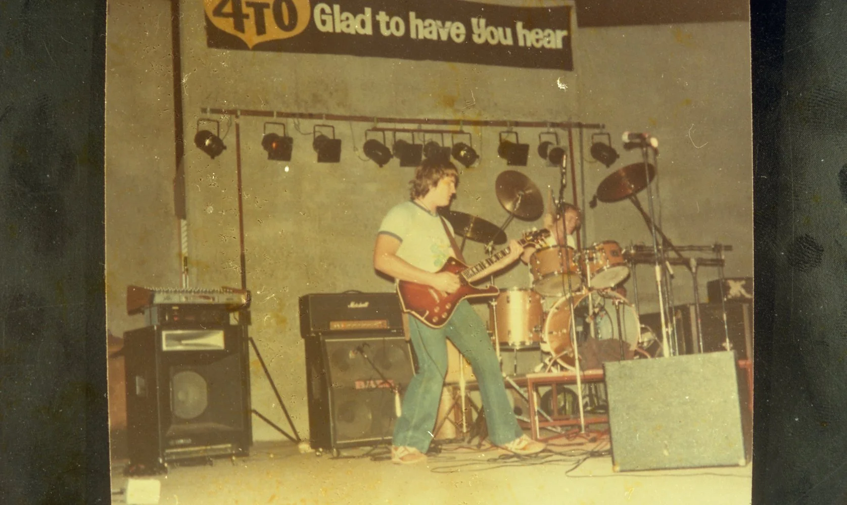 A band performs on stage with a guitarist and drummer. The stage has musical equipment and a sign that reads 'Glad to have You hear'. The photo appears vintage, with a yellowish tint.