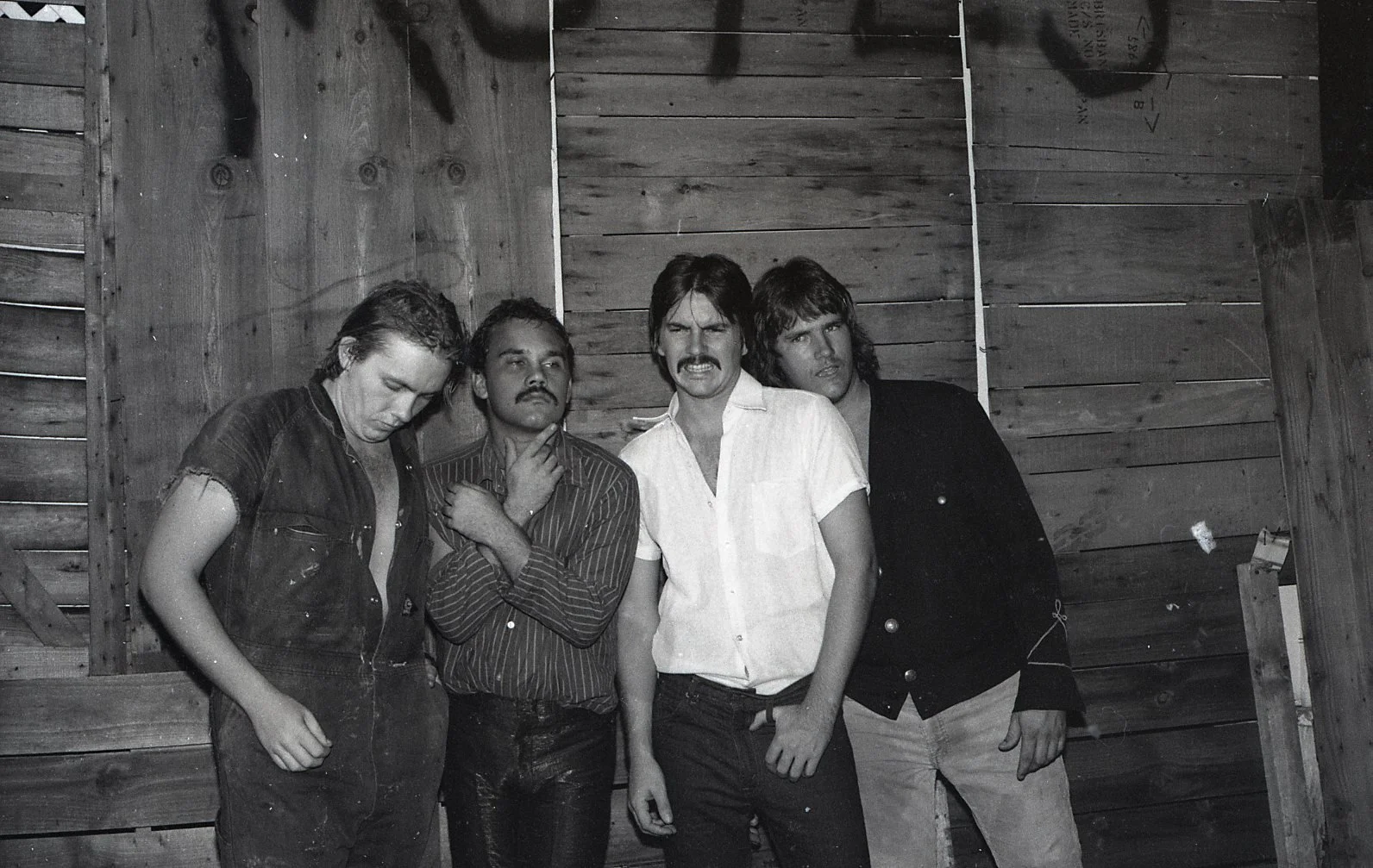 A black-and-white photo of four men standing against a wooden wall, with relaxed and expressive poses.