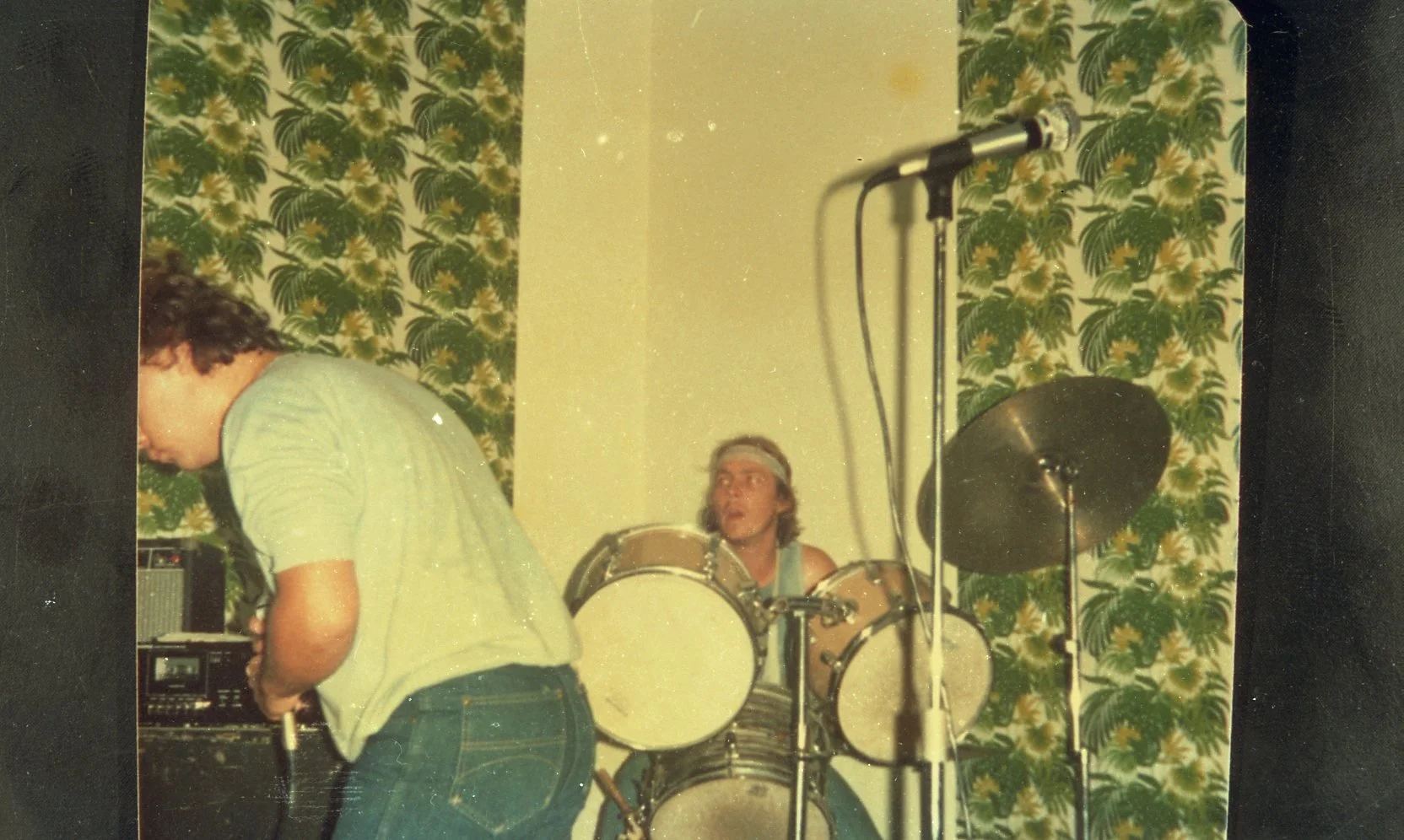 A band performing in front of a floral decorated wall, with a drummer in the center and a person adjusting equipment on the left.