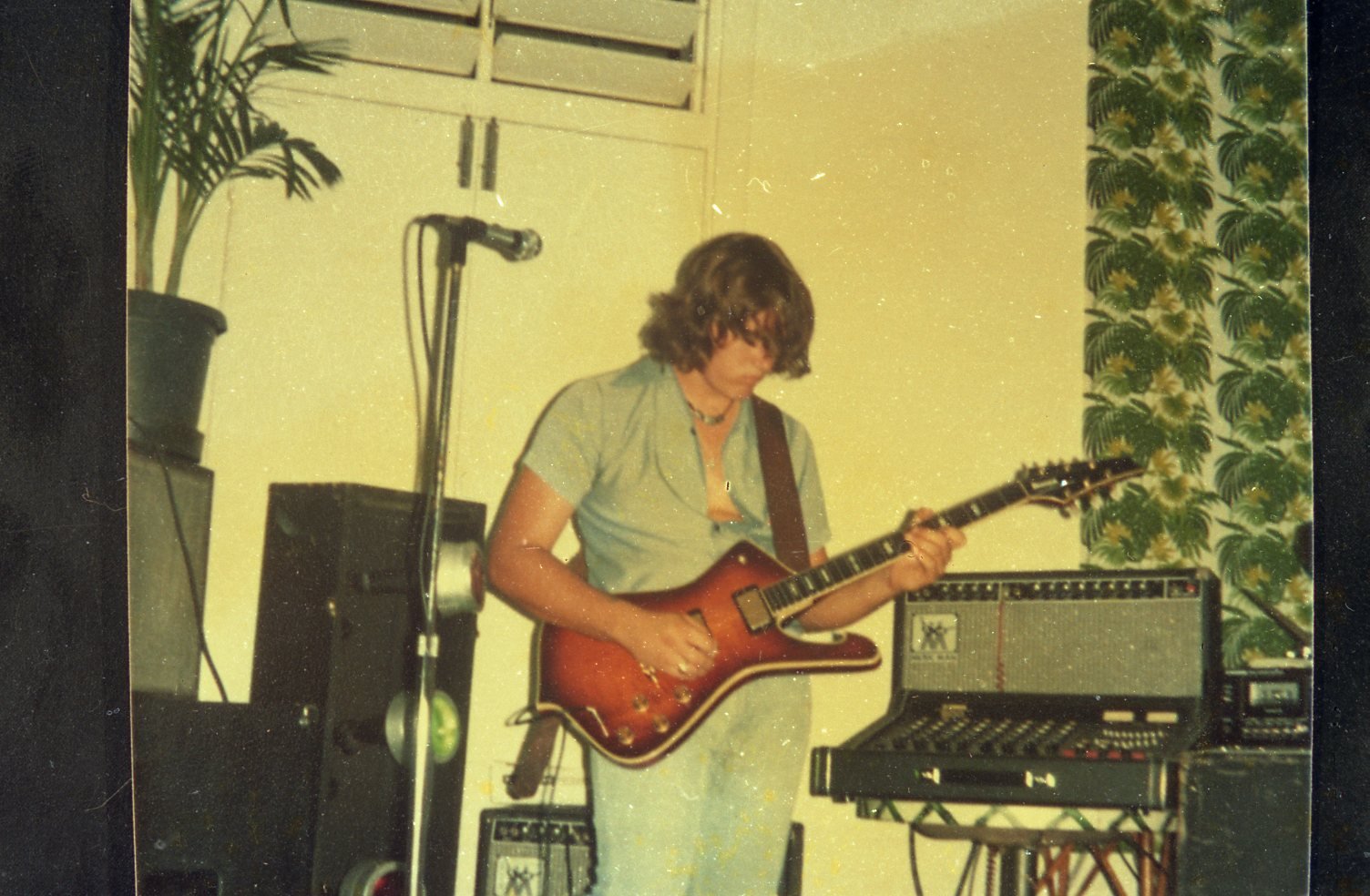 A person with shoulder-length curly hair playing an electric guitar in a room, surrounded by audio equipment, with a potted plant and patterned curtain in the background.
