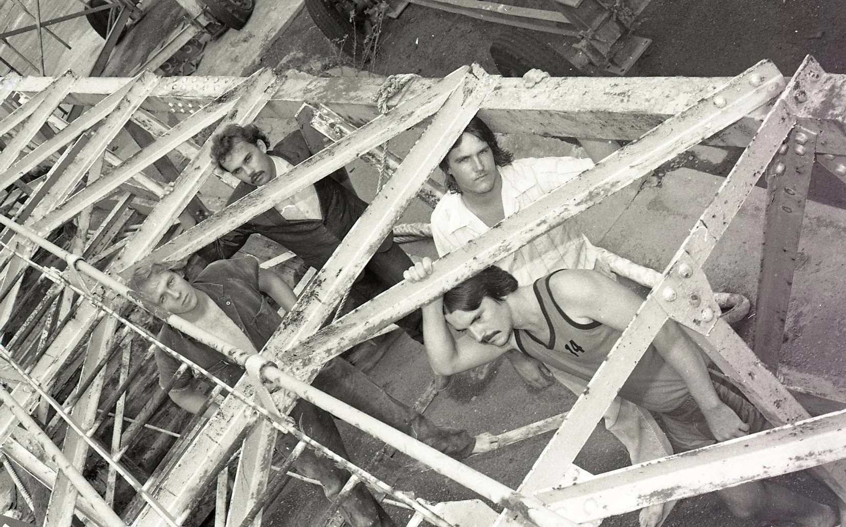 Four people are behind a metal structure, with three standing and one sitting, all with serious expressions in black and white photo.