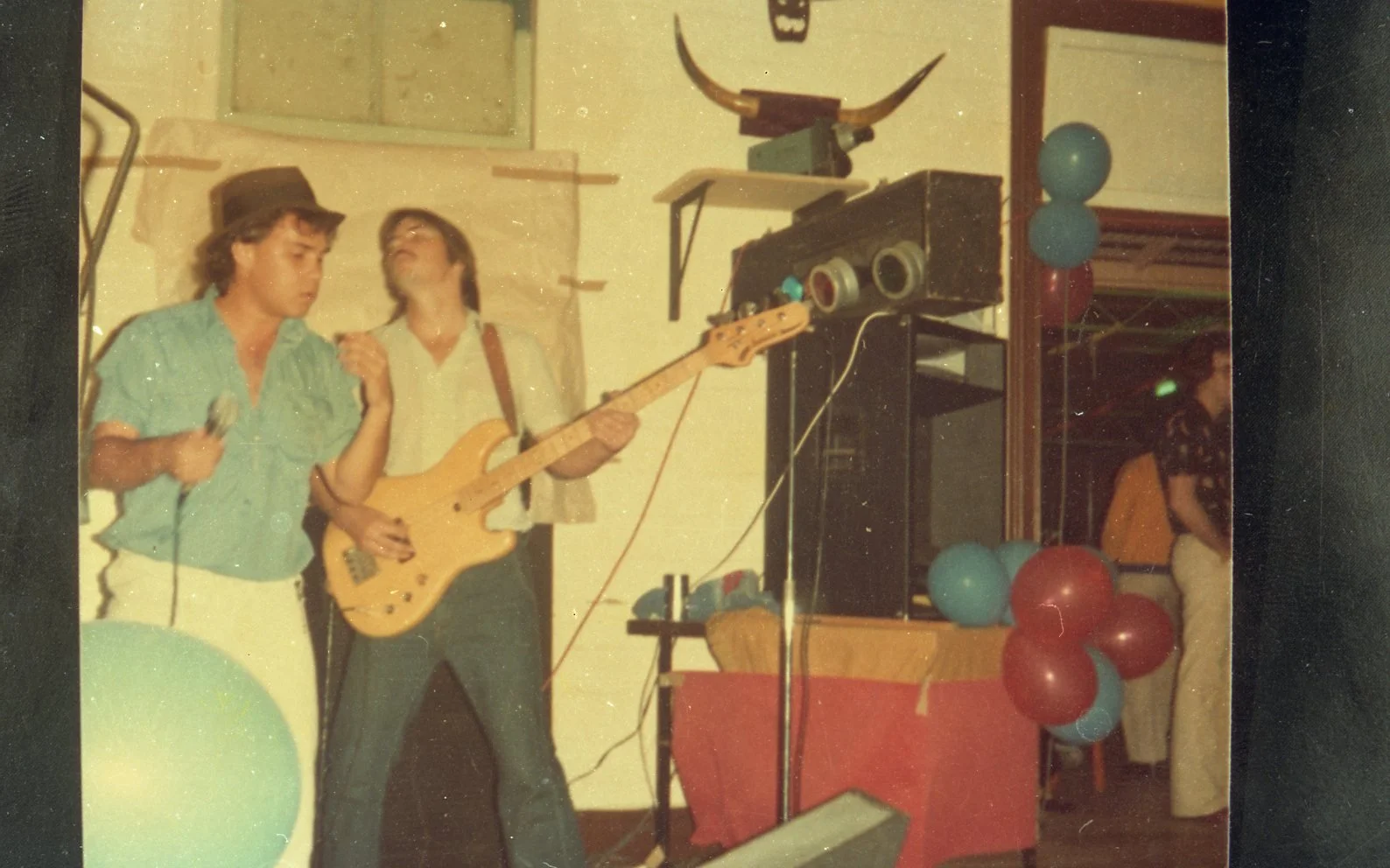 A vintage photo showing two young men performing at a party, one singing into a microphone and the other playing a bass guitar, with colorful balloons decorating the background.