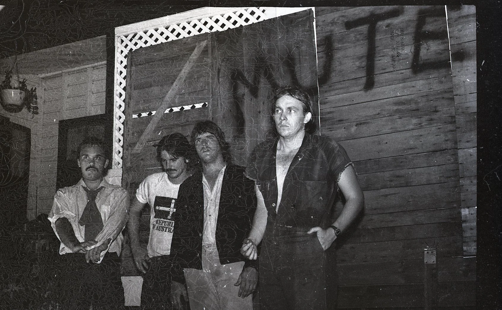 Black and white photo of four men standing indoors, with a wooden wall behind them. The man on the far left wears a white shirt and tie, appears serious. The second man wears a T-shirt with a design, has dark hair and a mustache. The third man wears 