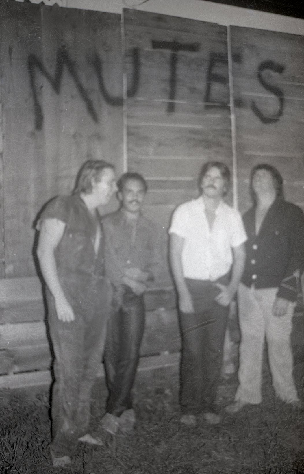 Four people standing in front of a wooden wall with the word 'MYTTS' spray painted above. The photo is black and white, and the individuals appear to be smiling and casually dressed.