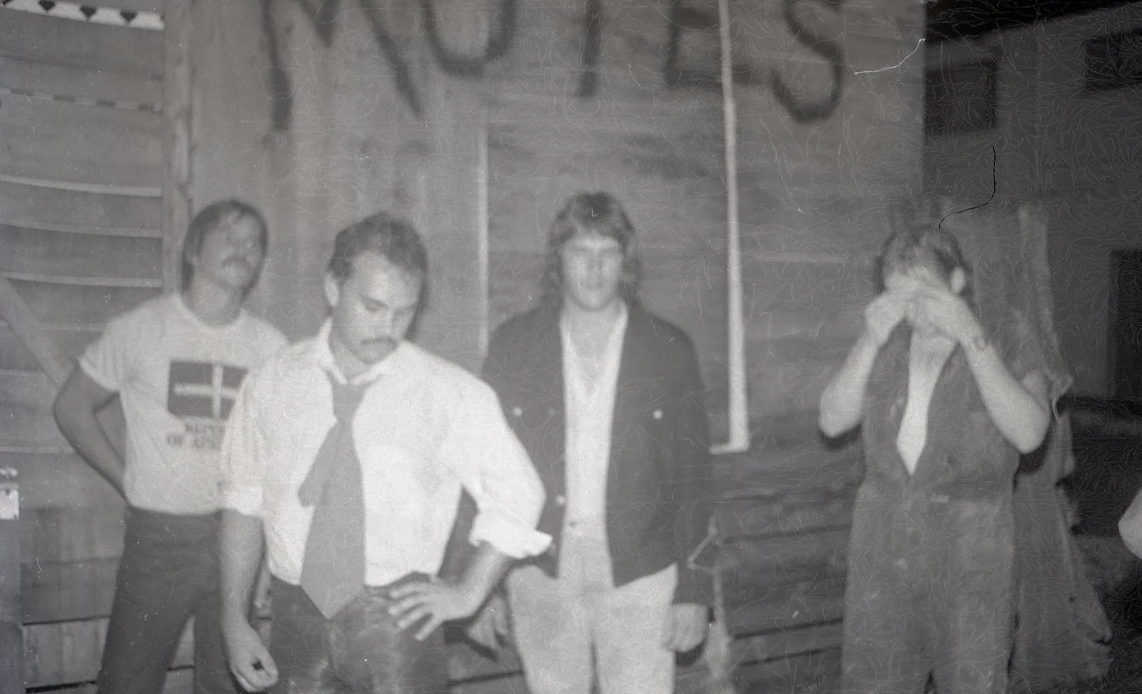 Black and white photo showing four people standing in a room with wooden walls. One person is holding a football, the others have different expressions, with the woman on the far right covering her face with her hands.