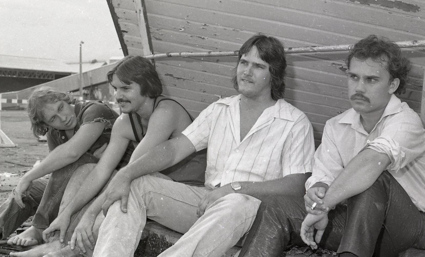 Four young men sitting and leaning against a weathered wall outdoors, with a fence and some structures visible in the background, in black and white.