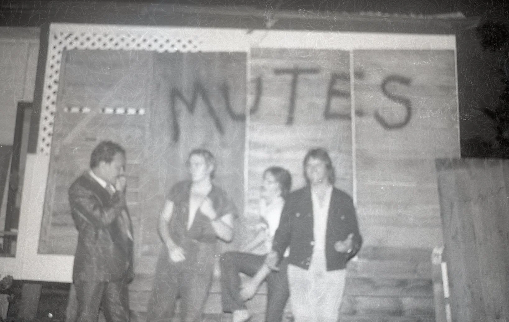 A black and white photo of four people standing in front of a wooden wall with the word 'NOTES' written above them. The man on the left is wearing a suit and appears to be adjusting or touching his face. The three women are standing in the middle and