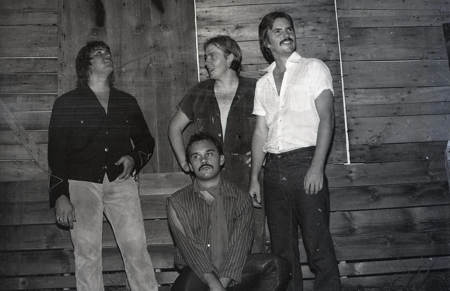 Black and white photograph of five people, four standing and one sitting, against a wooden wall. The standing individuals include three men and one woman, all dressed in casual or semi-casual clothing from the 1980s or early 1990s. The seated man has