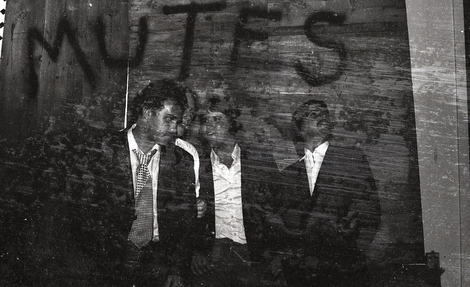 Three young men in formal attire standing in front of a graffiti-covered wooden wall, with partially visible graffiti text reading 'WTS'.