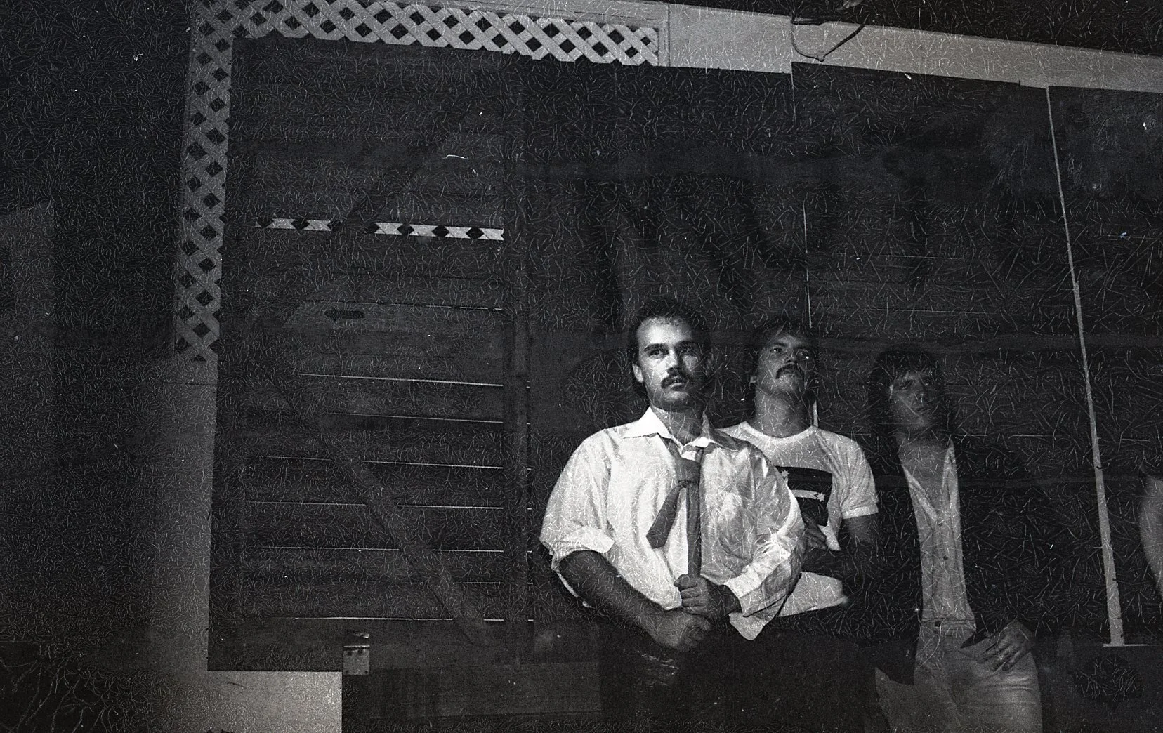 A black and white photo of three men reflected in a mirror, all wearing white shirts, with one holding a stick. The background includes patterned walls and a ventilation grate.