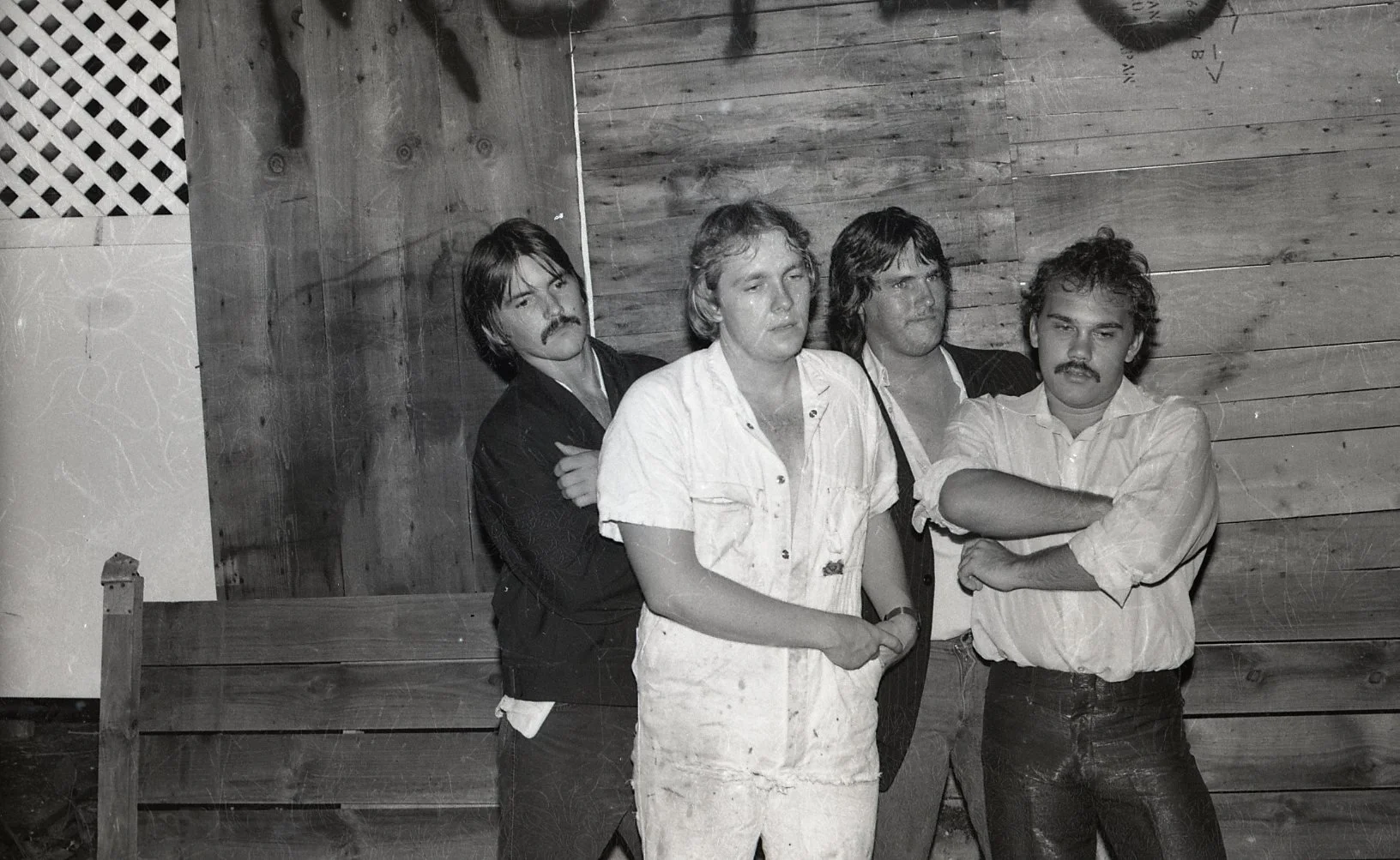 Black and white photo of four men and one woman standing with arms crossed or hanging, in front of a wooden wall. They have serious expressions, and all have hairstyles and clothing typical of the 1970s.