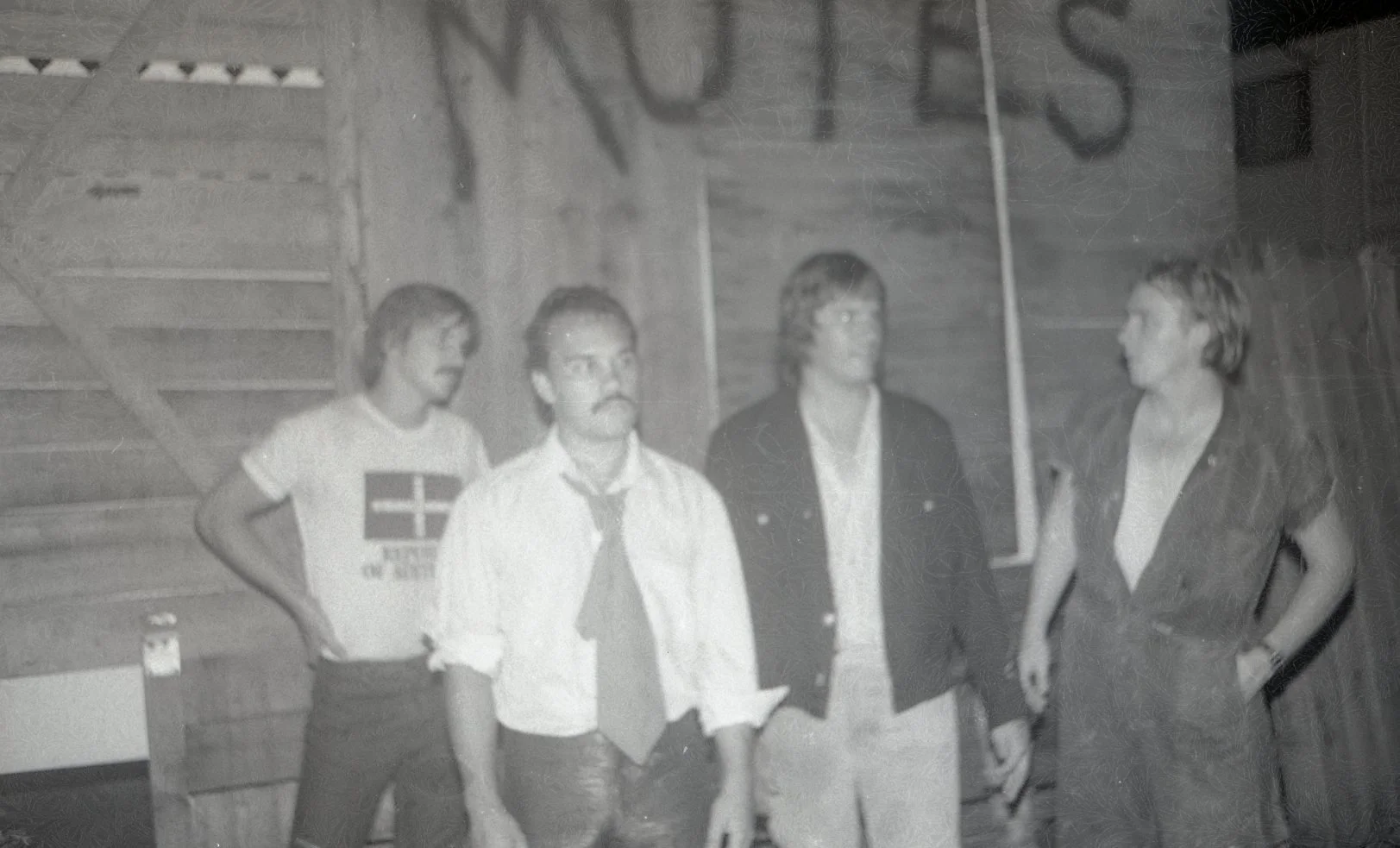 Four young people standing indoors near a wooden wall with stairs in the background, dressed casually, engaging in conversation or waiting.