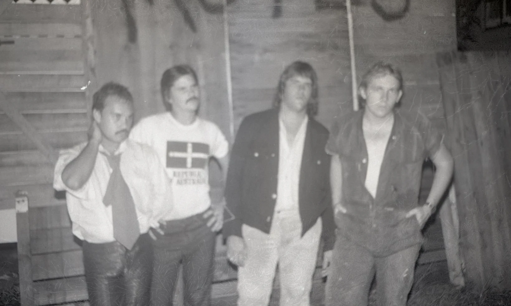 Four men standing indoors against a wooden wall, some with hands on hips or touching their heads, in black and white photo.