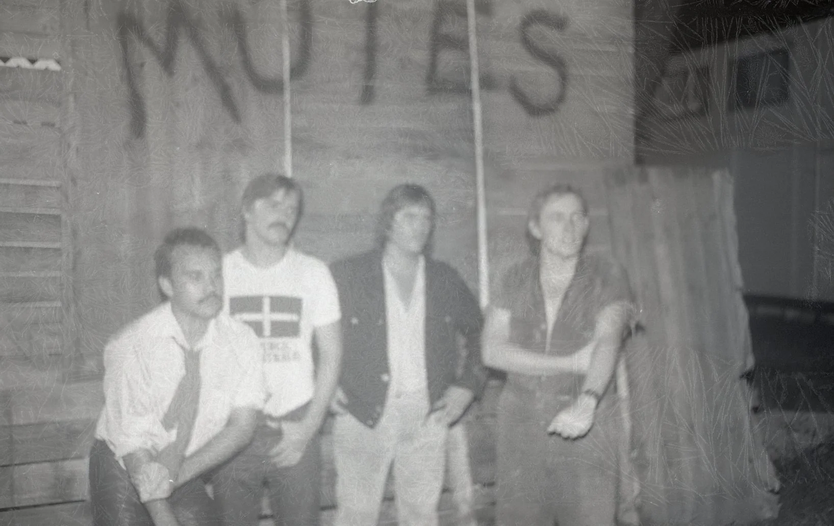 Four people, three men and one woman, standing together indoors against a wood-paneled wall. The woman is on the far right, wearing a dark jacket and light-colored pants. The man next to her, second from the right, wears glasses, a light shirt, and a