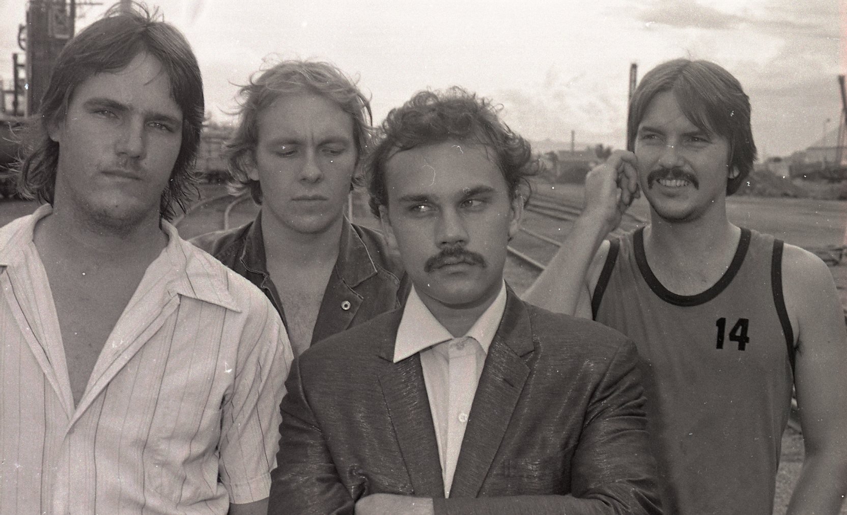Black and white photo of four men standing outdoors near train tracks, with industrial buildings in the background. The man on the left wears a striped shirt, the second man has curly hair and a serious expression, the third man with crossed arms has