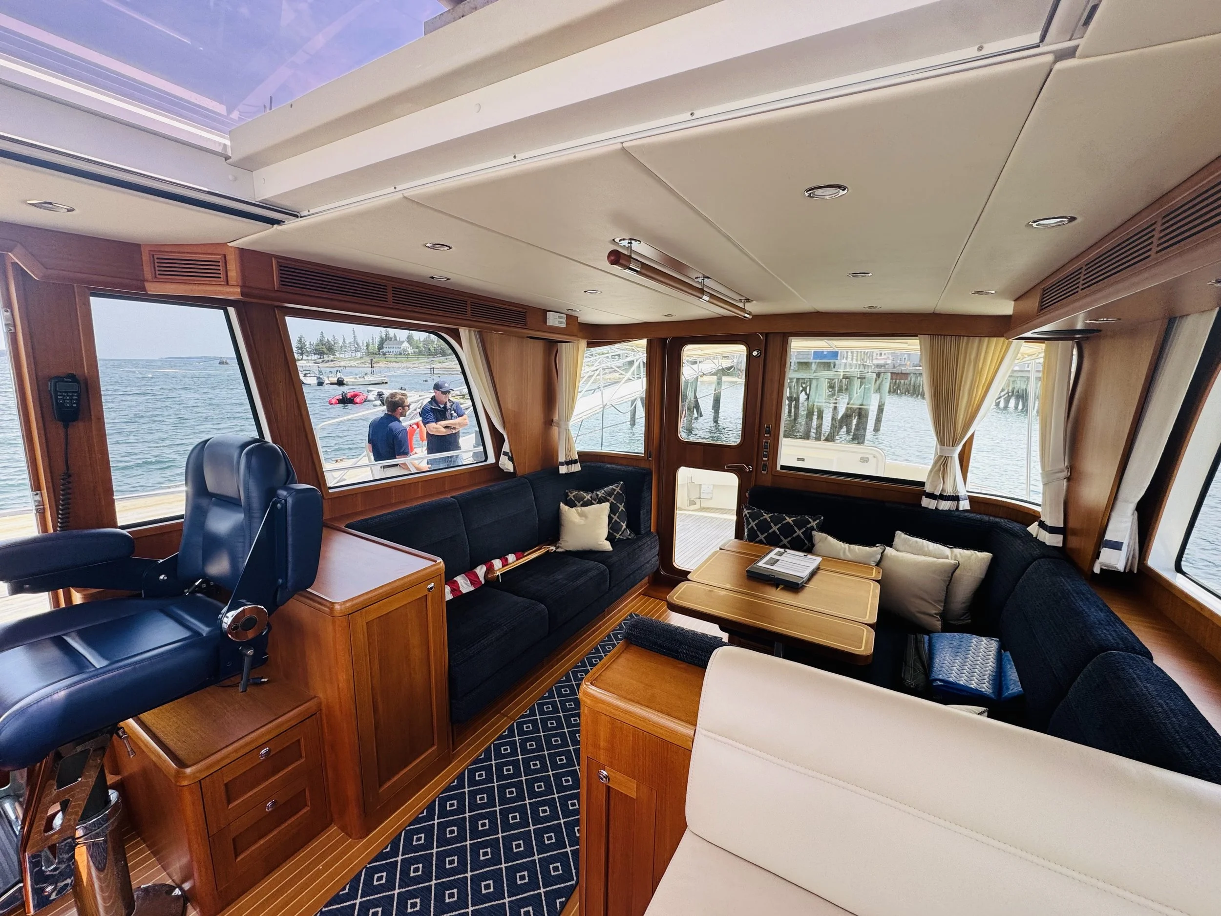 Interior of a boat with a seating area featuring dark blue and white cushions, wooden tables, and large windows showing a marina with boats and water.