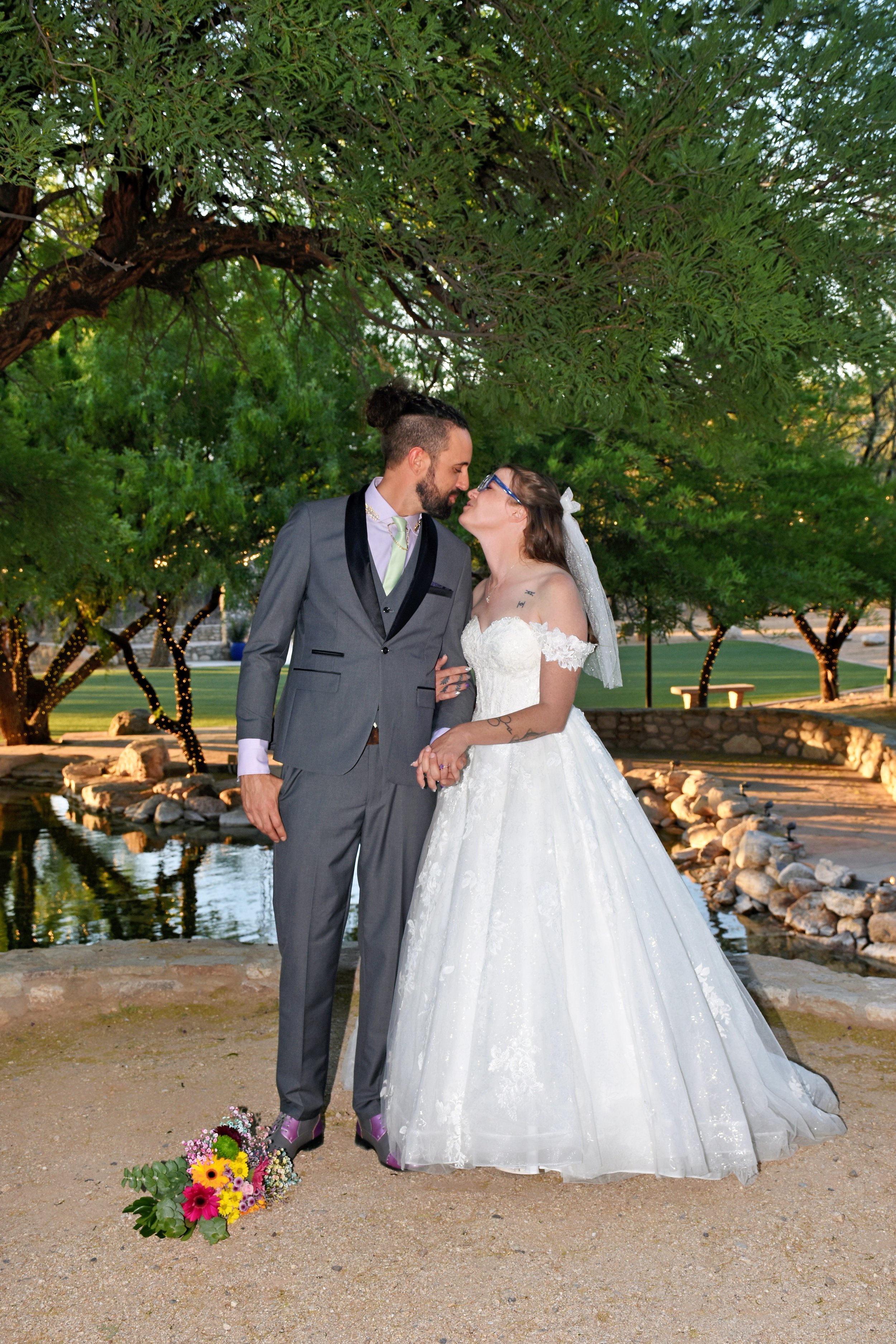 A couple in wedding attire standing by a small pond and rock formations in a park, surrounded by green trees and lit with string lights in the evening.