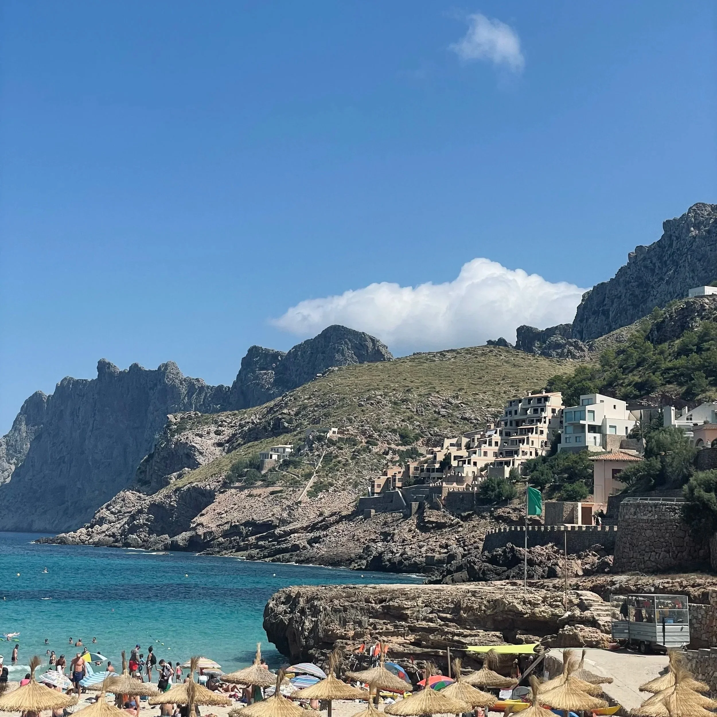 Beach scene with people swimming and sunbathing beneath straw umbrellas, rocky coastline, clear teal water, and hillside buildings with mountains in the background.