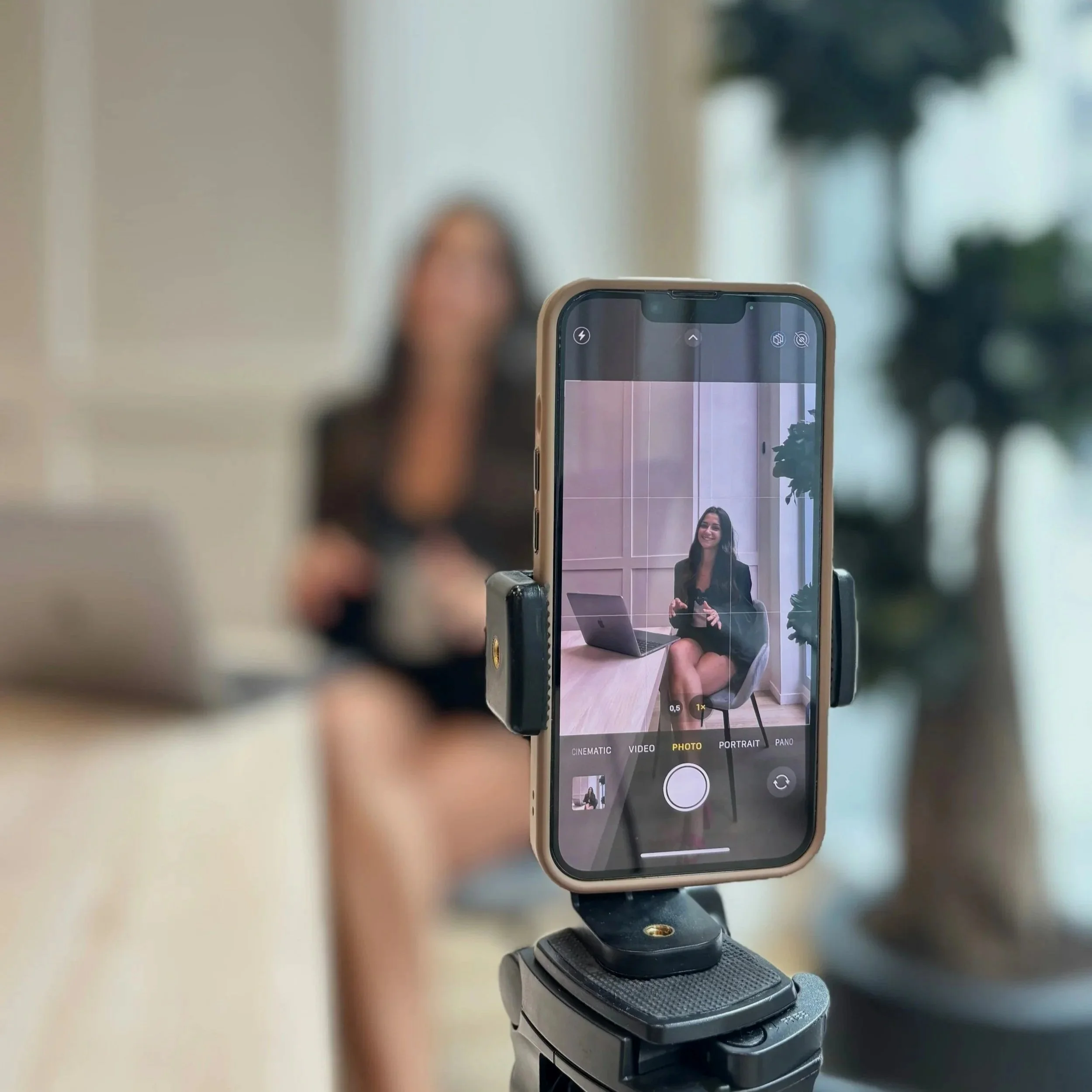 Close-up of a smartphone on a tripod, taking a selfie of a woman with long dark hair, sitting at a desk with a laptop, in a room with light-colored walls and a large window in the background.