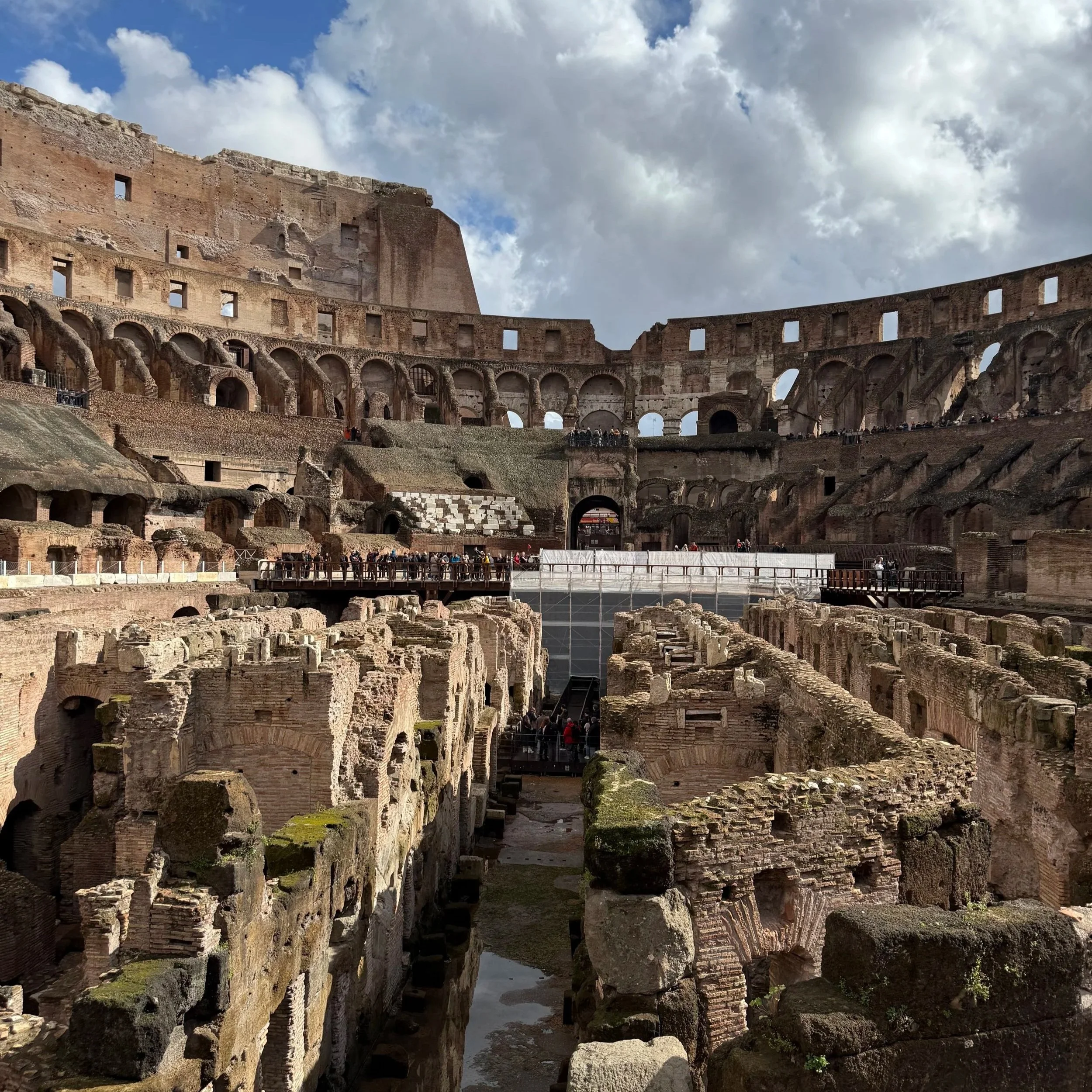 Interior of the ancient Roman Colosseum with partially ruined stone walls, arches, and spectator areas under a partly cloudy sky.