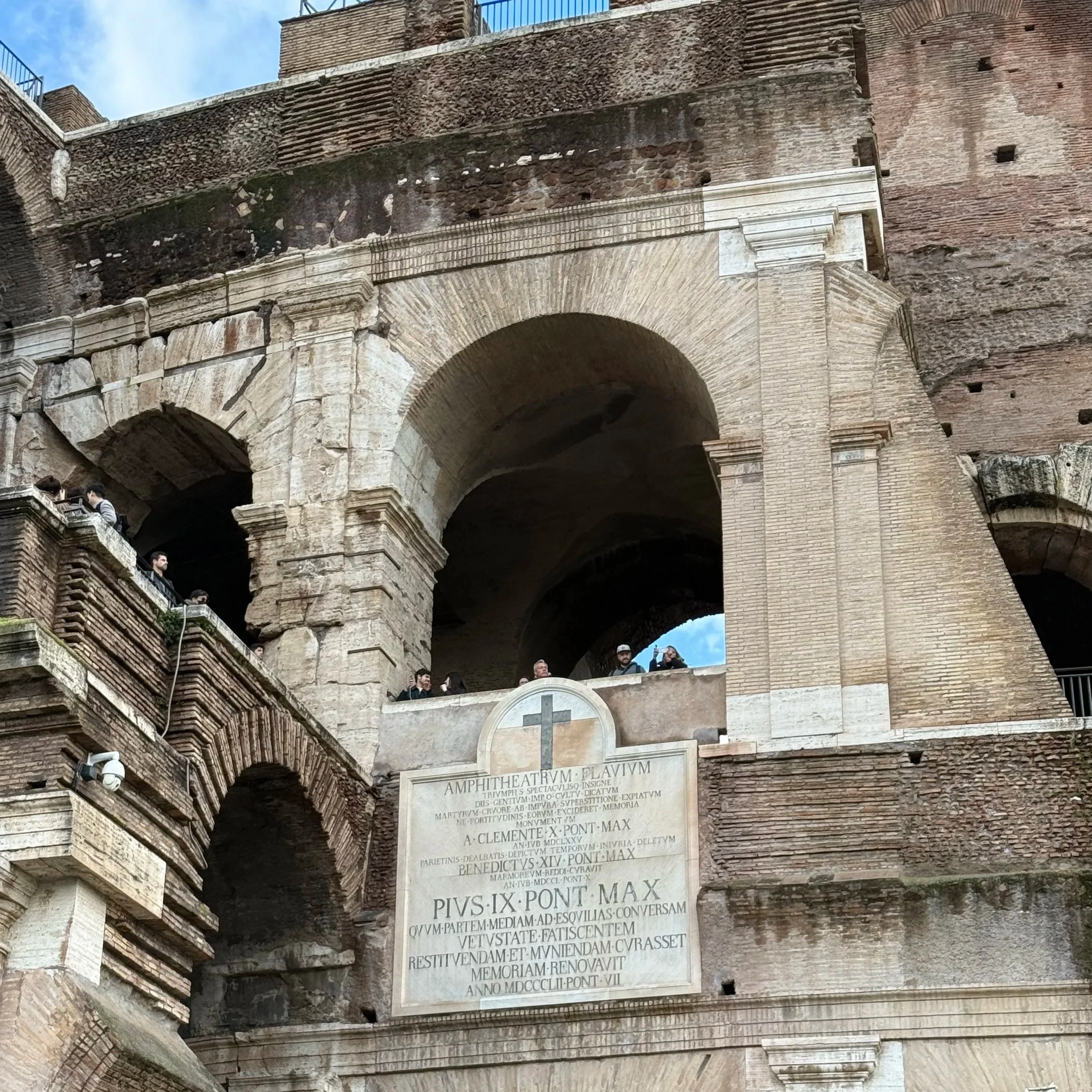 Close-up of ancient Roman ruins with stone arches, weathered brickwork, and Latin inscriptions on a marble plaque, with people exploring the site.