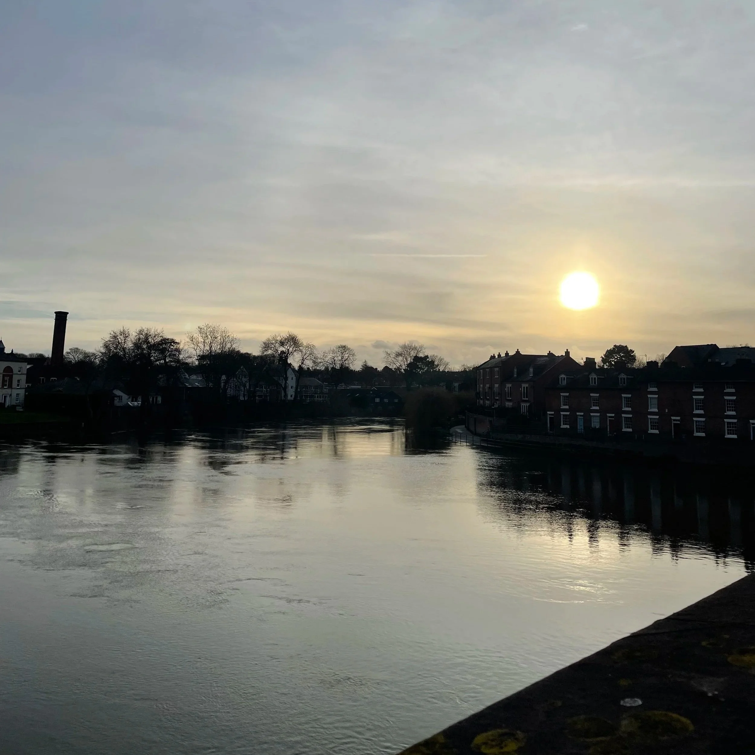 Sun setting over a calm river with trees and houses along the bank, and a chimney in the town.