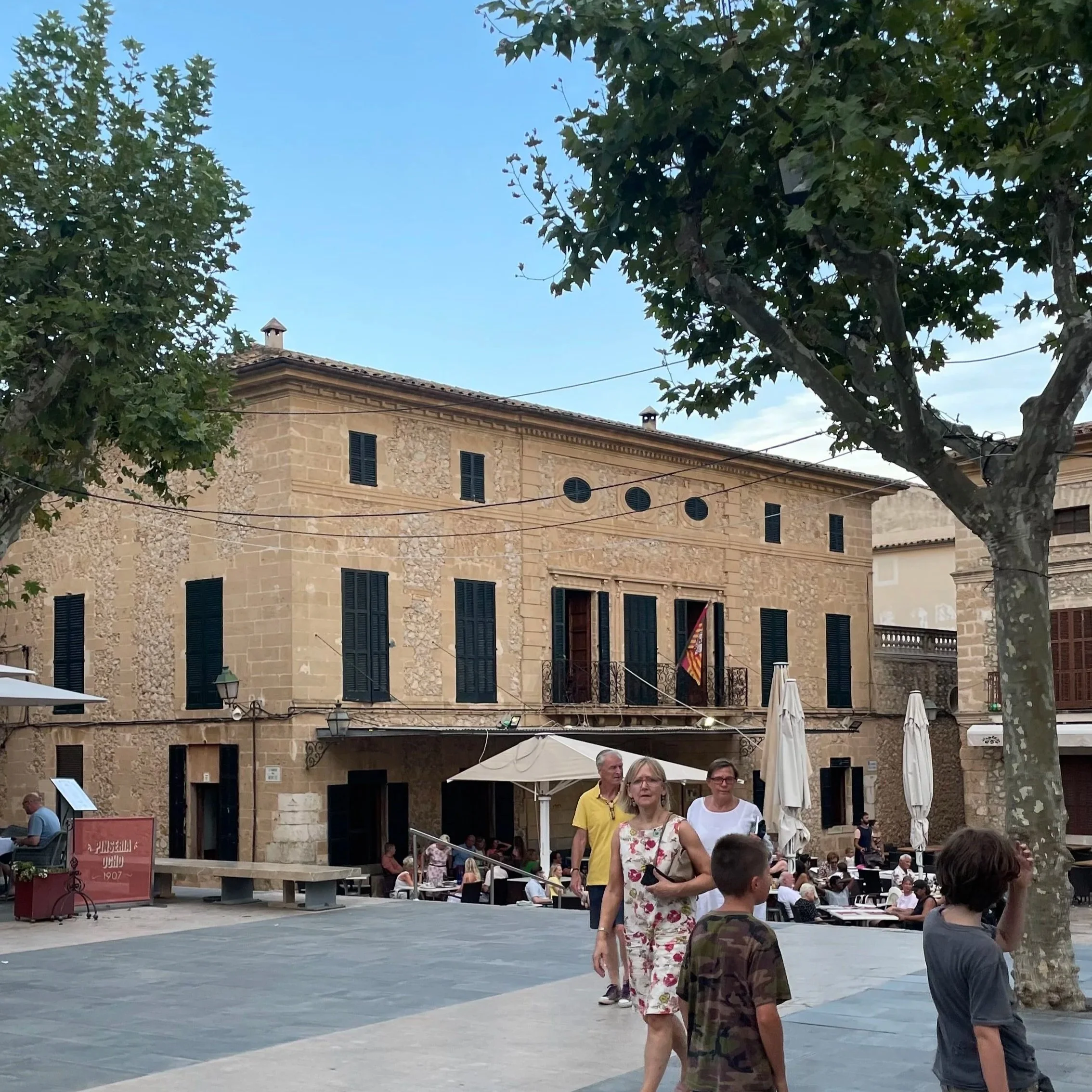 People walking and sitting in a courtyard surrounded by tall stone buildings with green shutters and trees, under a blue sky.