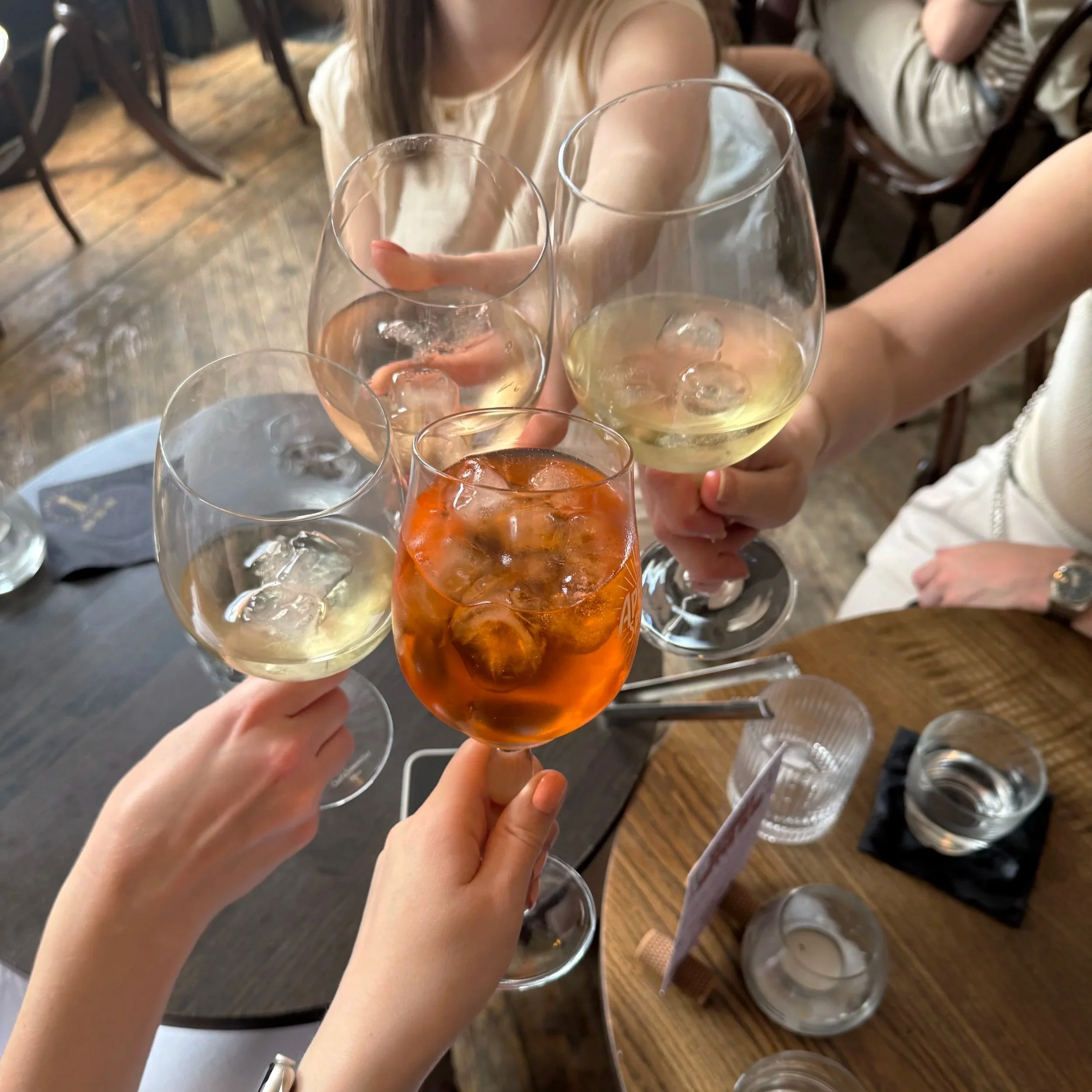 Close-up of people clinking wine glasses filled with white wine, ice, and orange-colored drink in a restaurant with wooden floors and chairs.
