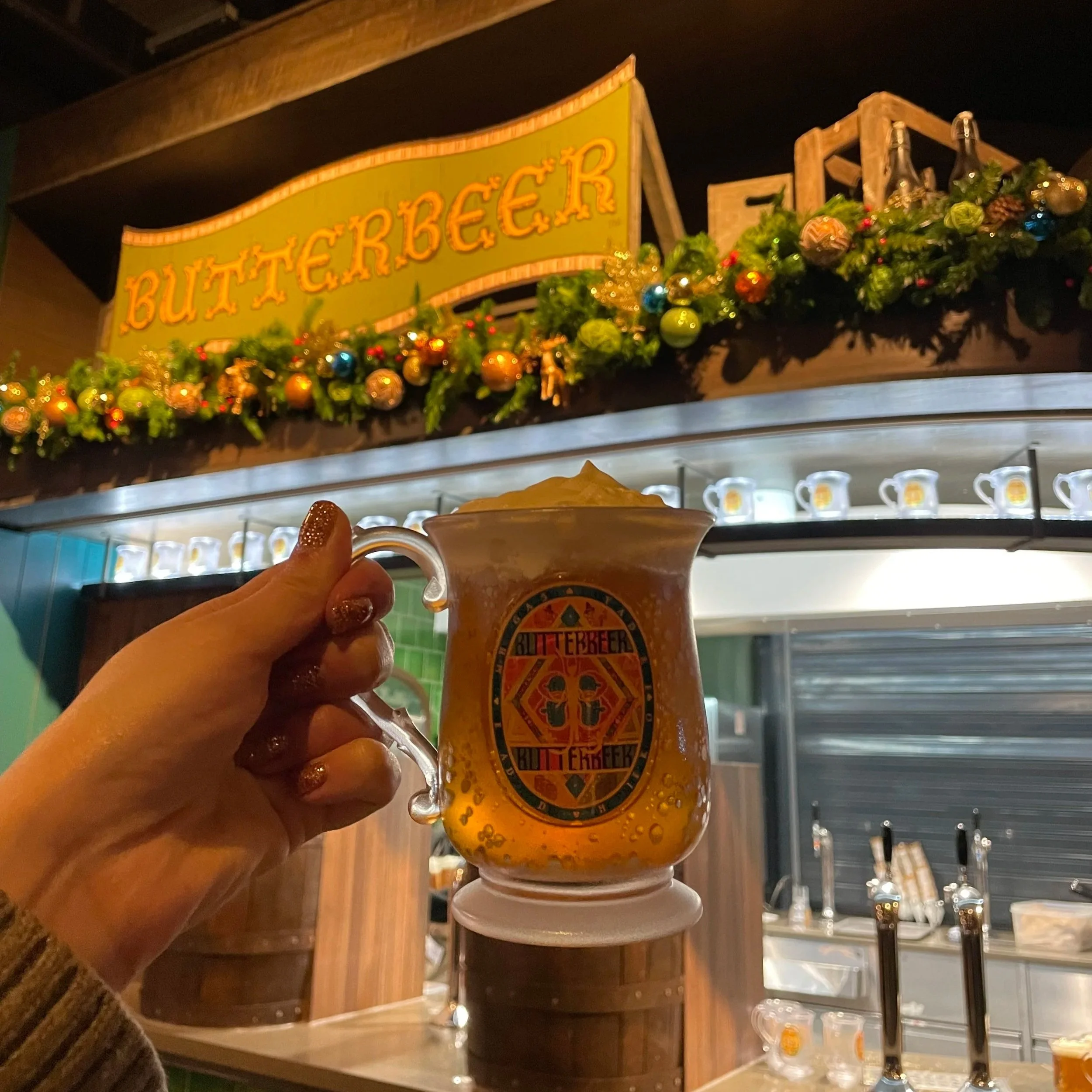 A person holding a mug with a beer logo, in front of a bar with a decorated sign that says 'BUTTERBEER' and shelves of beer bottles, with festive holiday decorations including green foliage, ornaments, and lights, in a dimly lit setting.