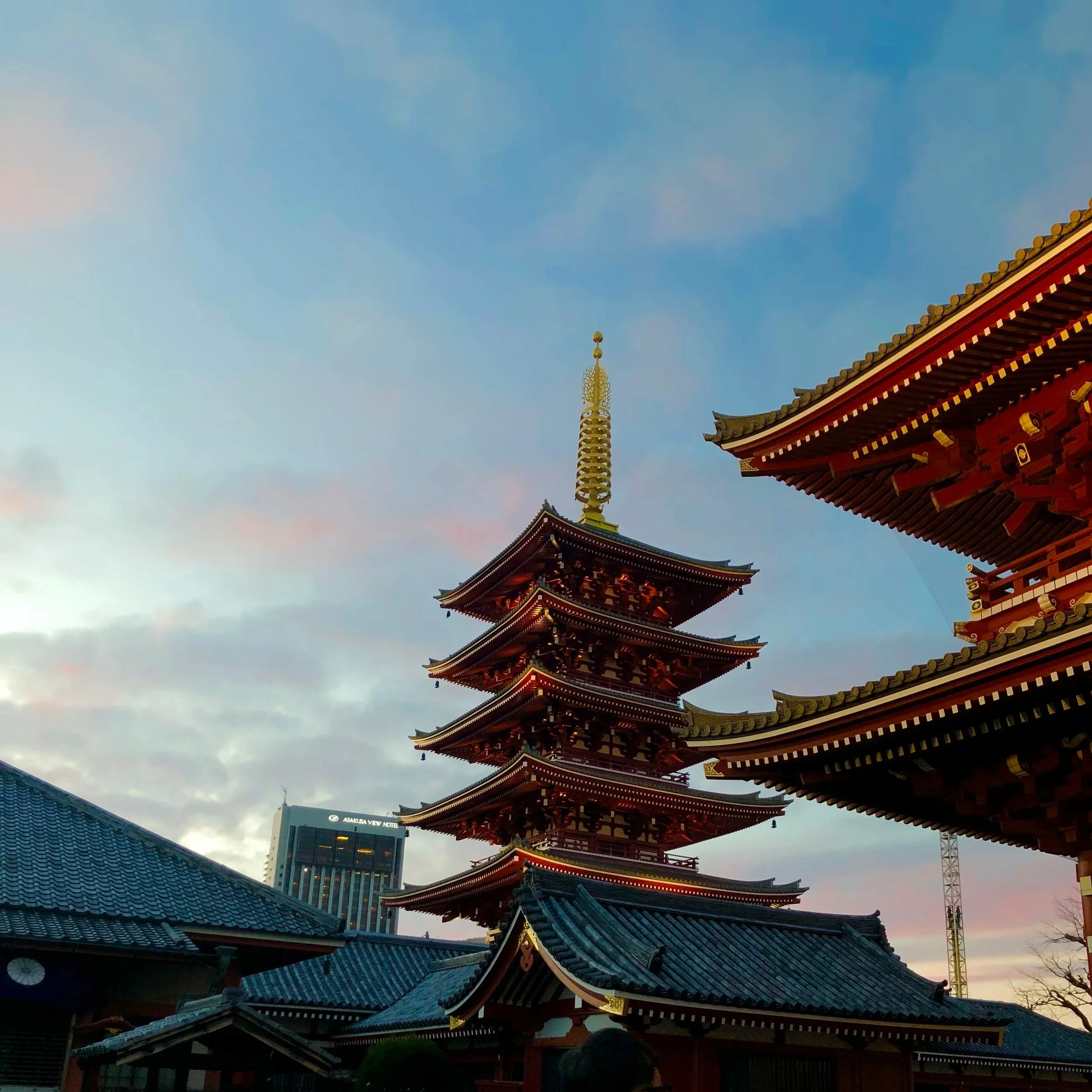 Traditional Japanese pagoda with red and white colors and tiered roofs, set against a sky during dusk with buildings and a crane visible in the background.