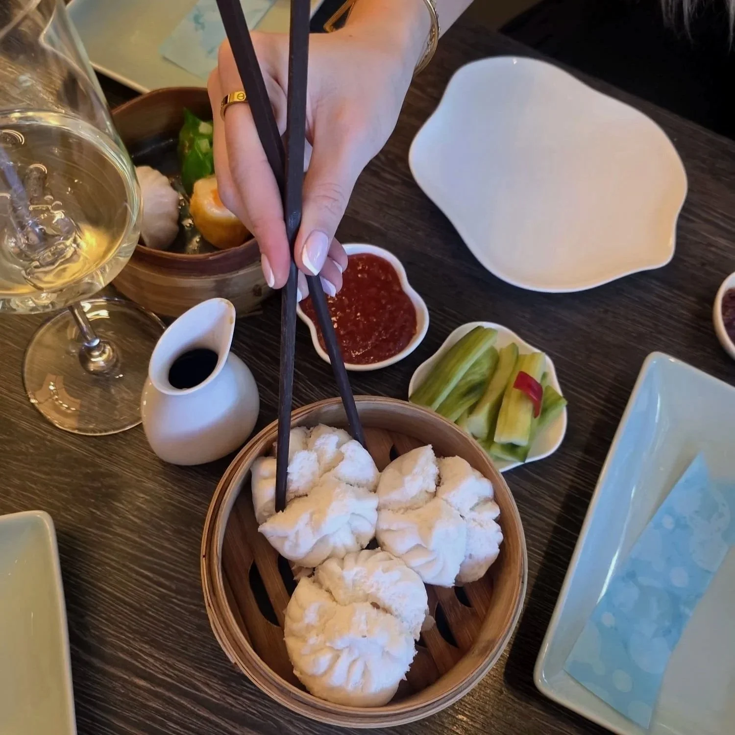 A person using chopsticks to pick up steamed buns at a dining table with various dipping sauces, vegetables, and a glass of white wine.