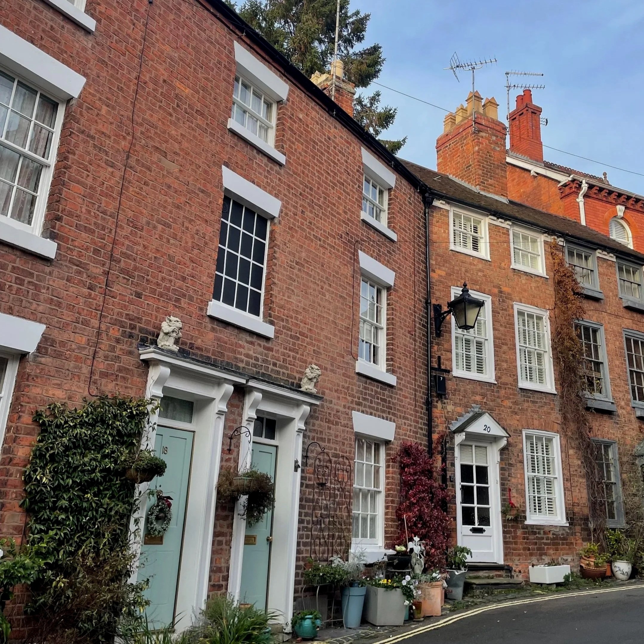 A row of traditional brick townhouses with colorful front doors, flower arrangements, and plants outside on a quiet street under a clear blue sky.