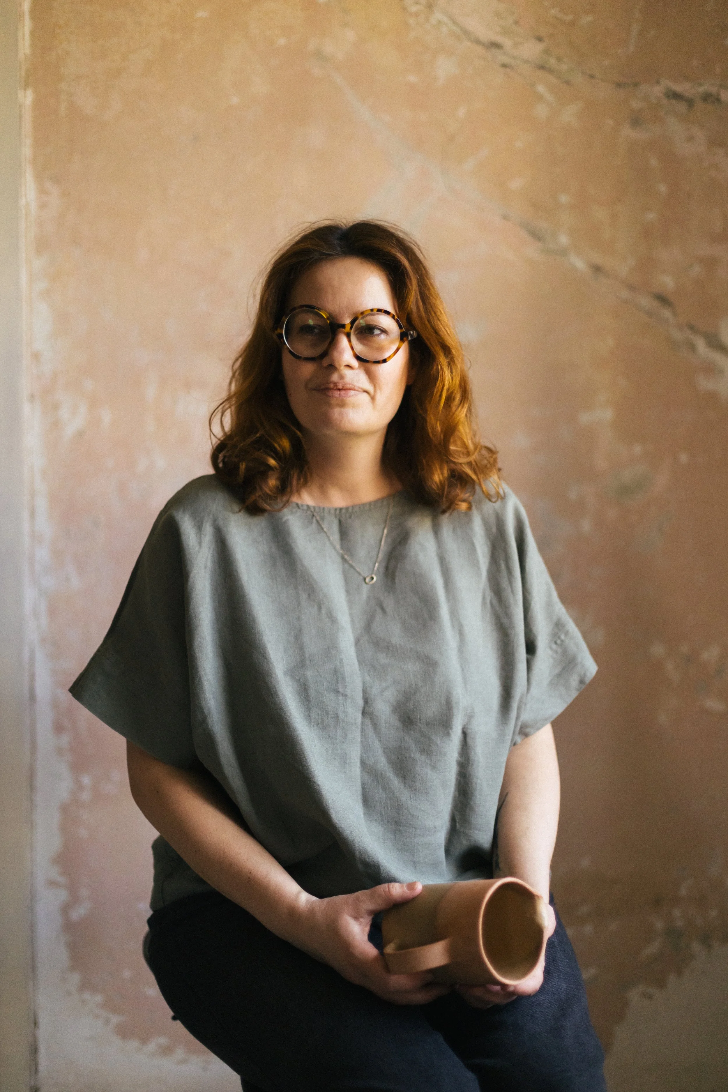 A woman with red, wavy hair and glasses holding a tan ceramic mug, sitting against a distressed pink and beige wall.