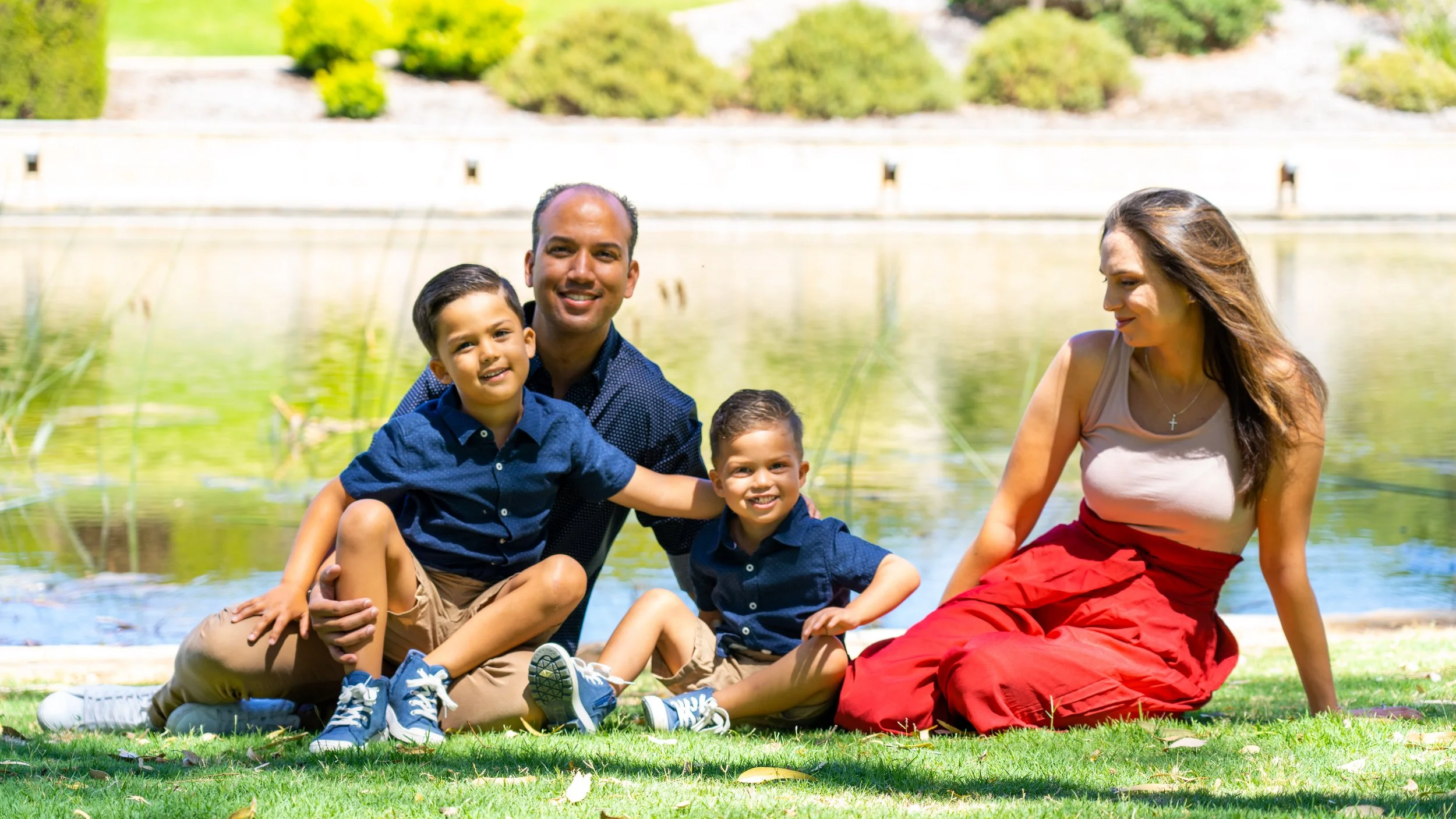 A family of five, including two young boys, a man, and a woman, sitting by a pond in a park on a sunny day. They are smiling and enjoying the outdoors.