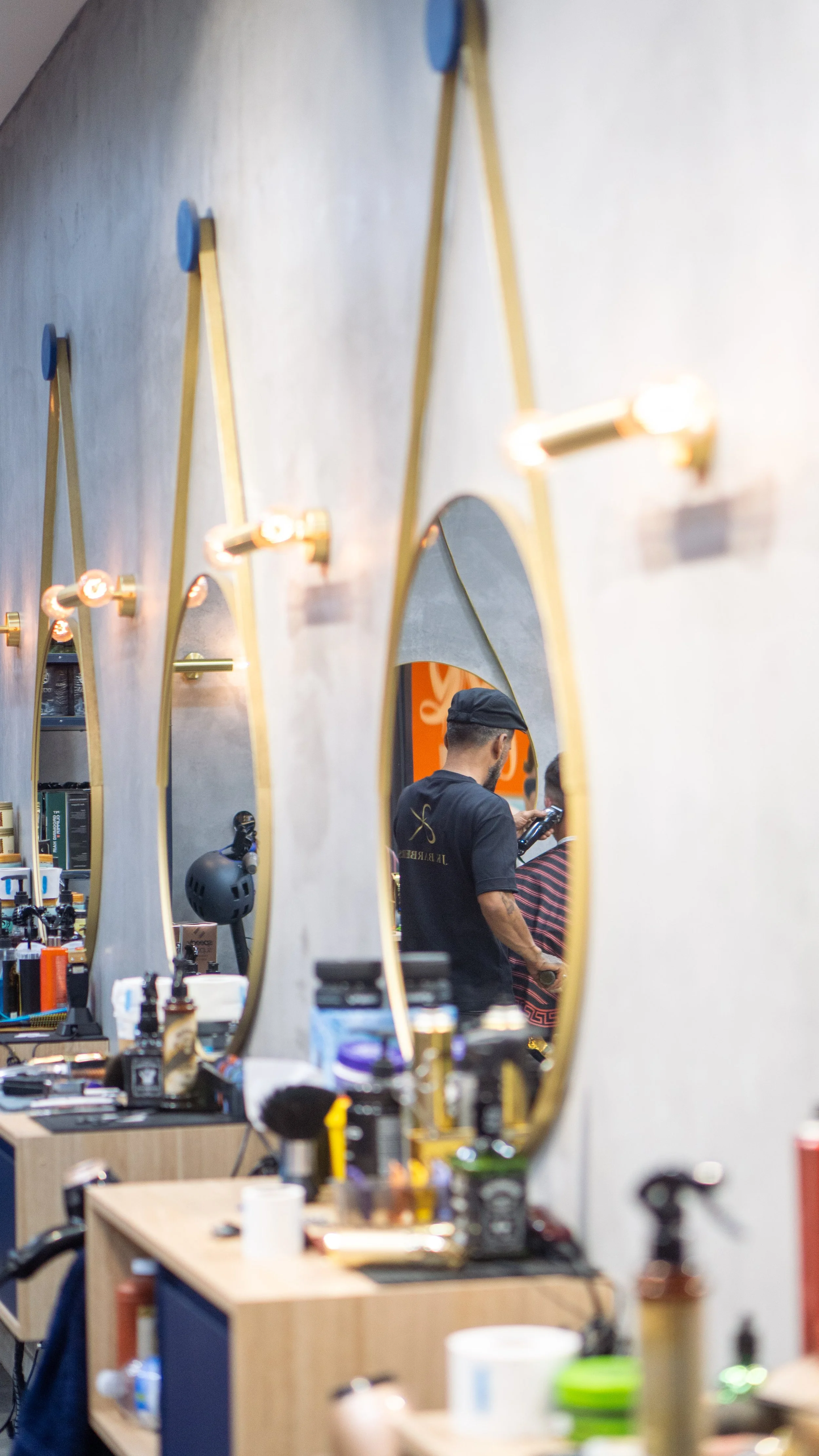 Inside a barbershop with a mirror showing a barber cutting a customer's hair, various grooming products on the counter, and decorative mirrors on the wall.