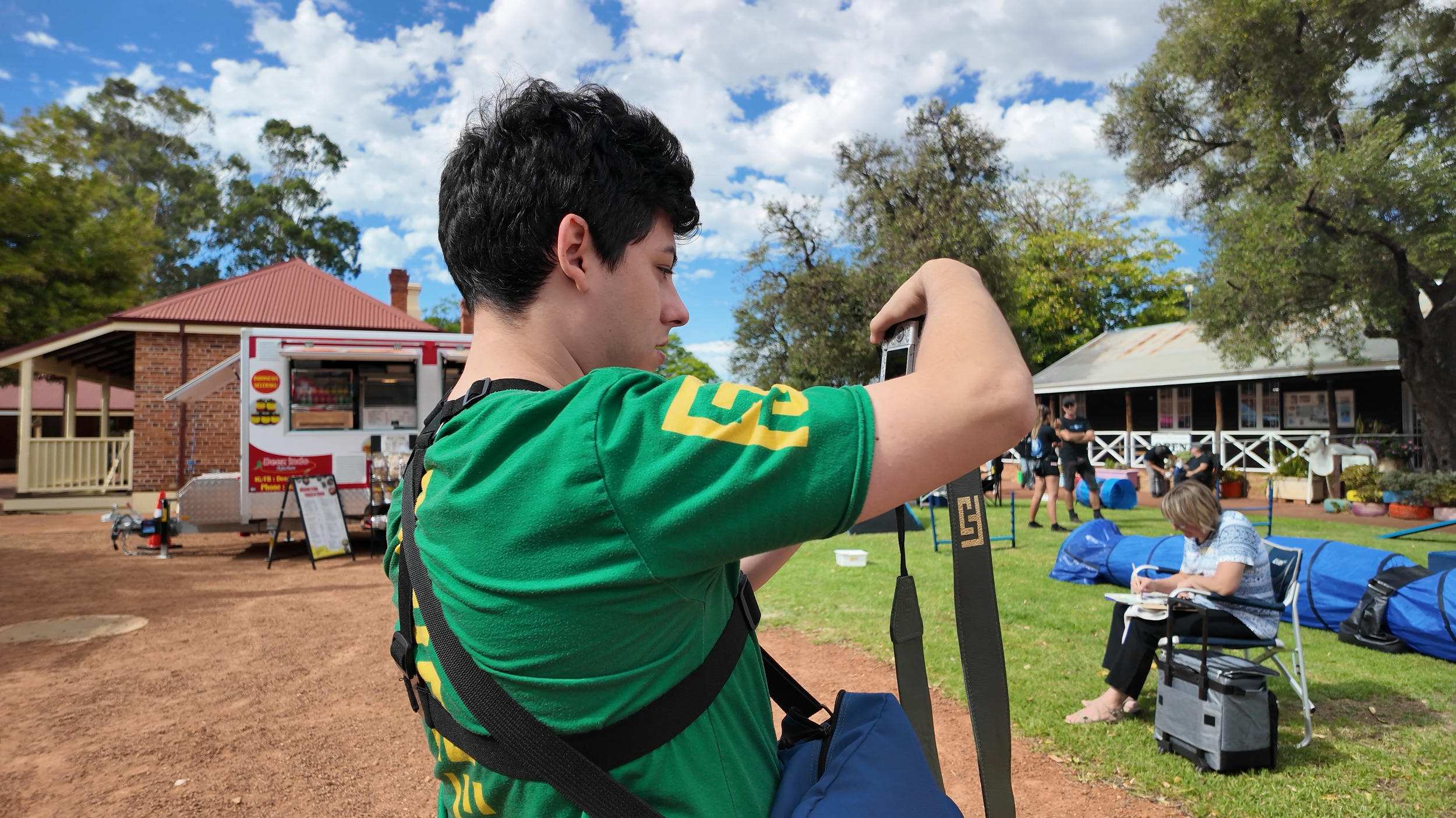A young man with dark curly hair wearing a green shirt and a camera strap around his neck is adjusting a camera or camera strap while outdoors at a park or fair. In the background, there are food trucks, people, and a building with a red roof, trees, and a partly cloudy sky.