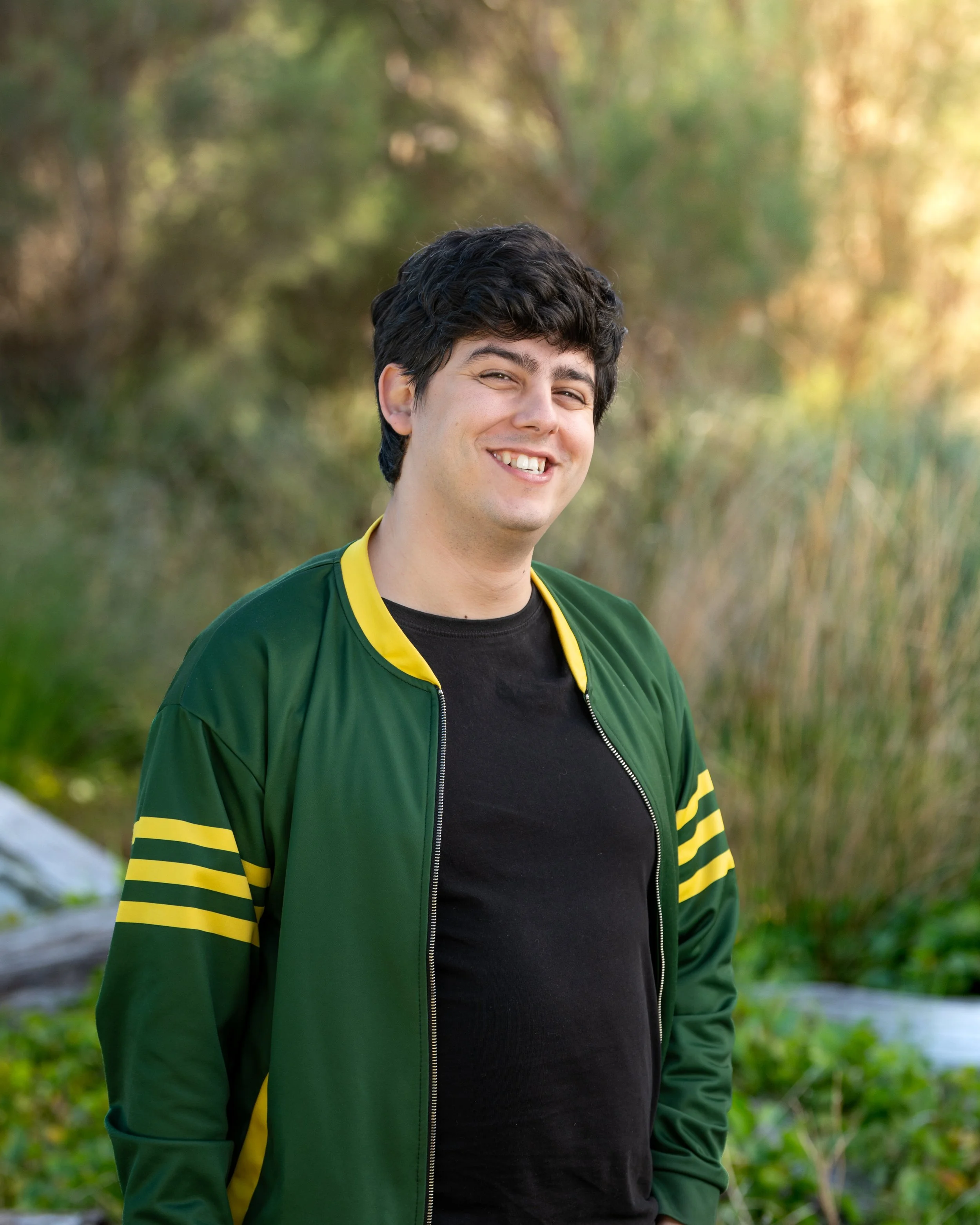 A young man with dark, curly hair smiling outdoors, wearing a green jacket with yellow accents and a black t-shirt, standing in a natural setting with blurred green and brown foliage in the background.