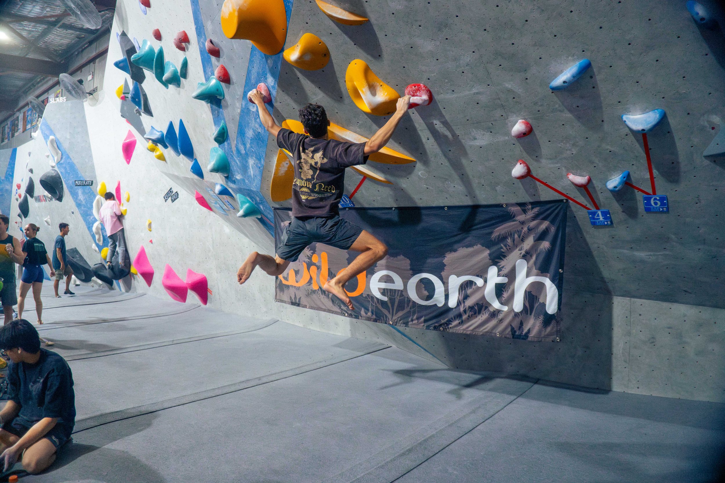Indoor rock climbing gym where a person is ascending a colorful climbing wall with holds of various shapes and colors, and others are resting or preparing nearby.