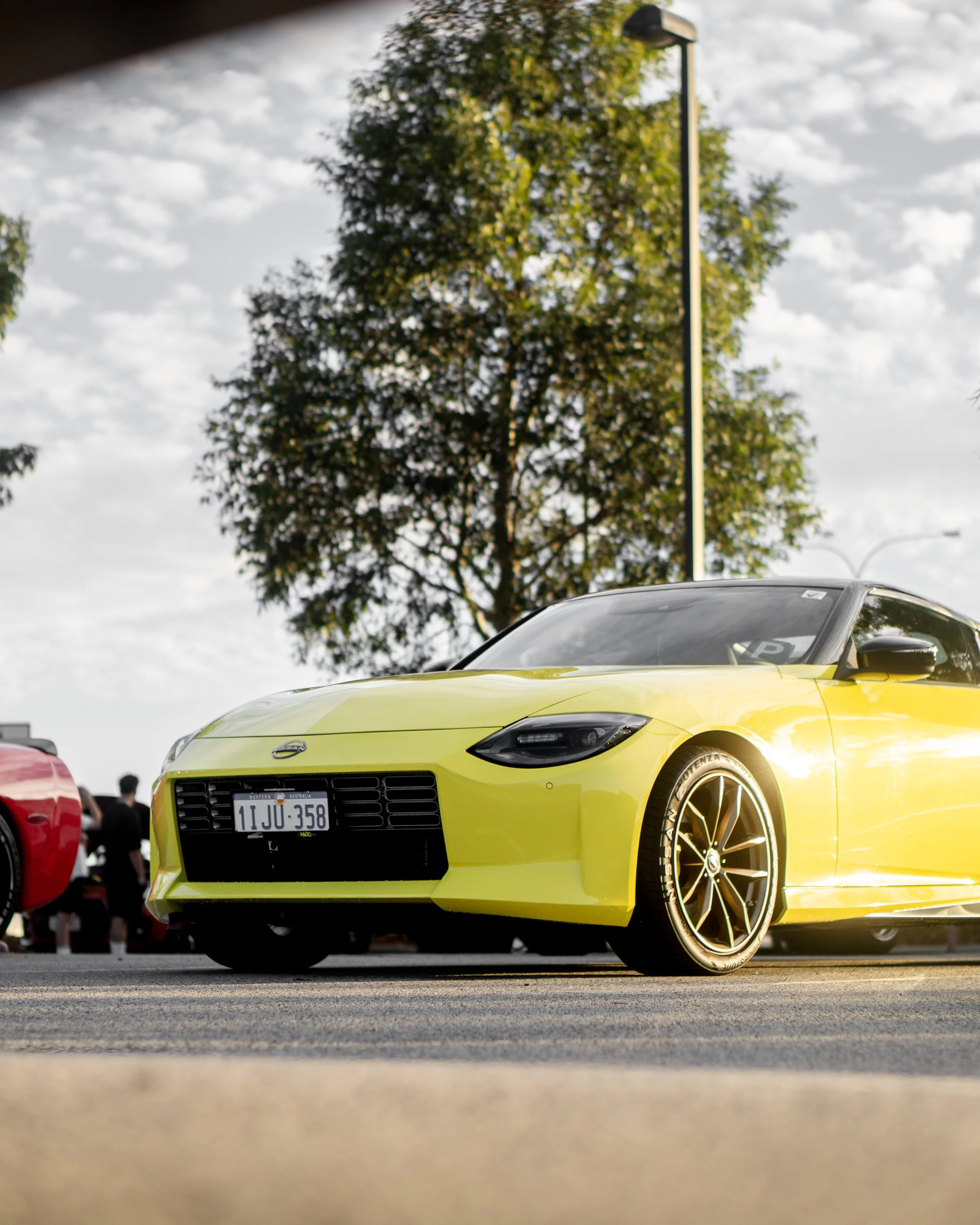 Bright yellow sports car with black rims parked outdoors at a car show, with a large tree, cloudy sky, and people in the background.