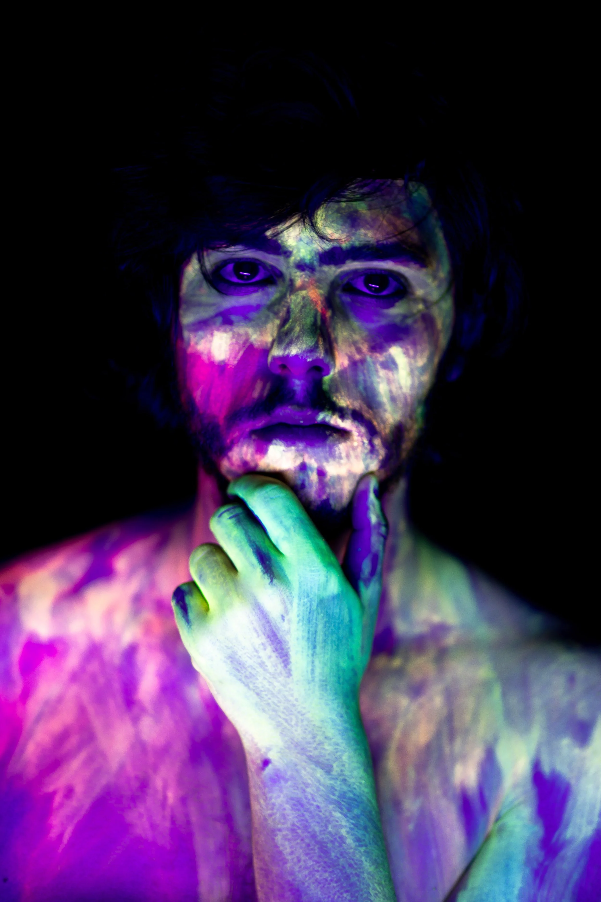 A person with dark hair and a beard, illuminated by multicolored lights, with their hand on their chin, looking contemplative. The background is dark.
