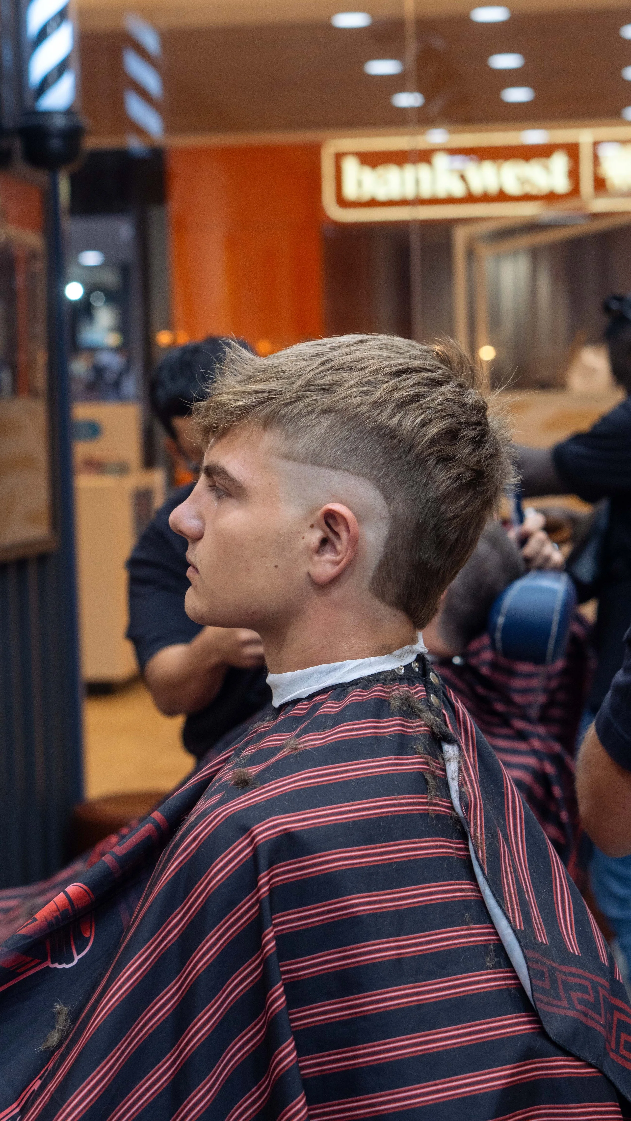 A young man with a hairstyle featuring shaved side and back, textured top, and a stripe pattern at the side, sits in a barbershop with a striped barber cape. Other customers and the barbershop interior are visible in the background.