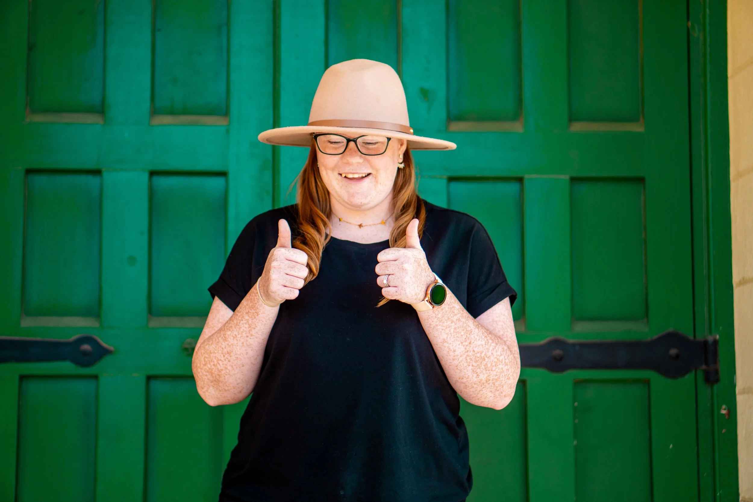 A woman with freckles, red hair, glasses, and a broad smile, wearing a beige wide-brimmed hat and black t-shirt, giving two thumbs up in front of a green wooden door.