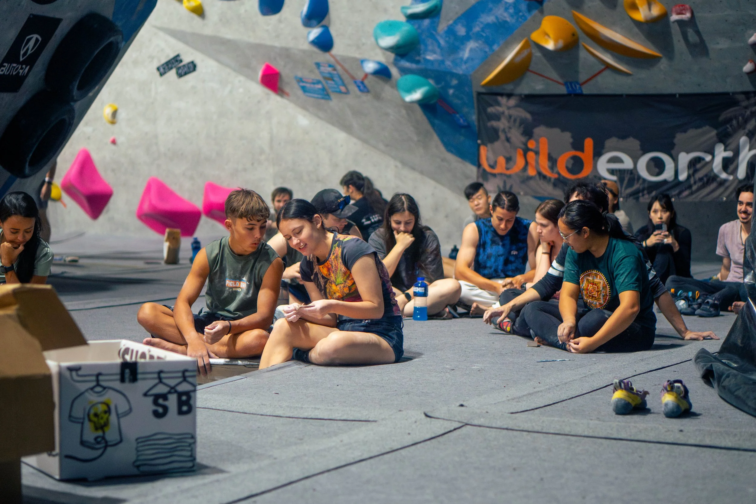A group of people sitting on the floor in front of an indoor climbing wall, possibly during a training or break, with colorful climbing holds and a banner that says 'Wilderness' in the background.