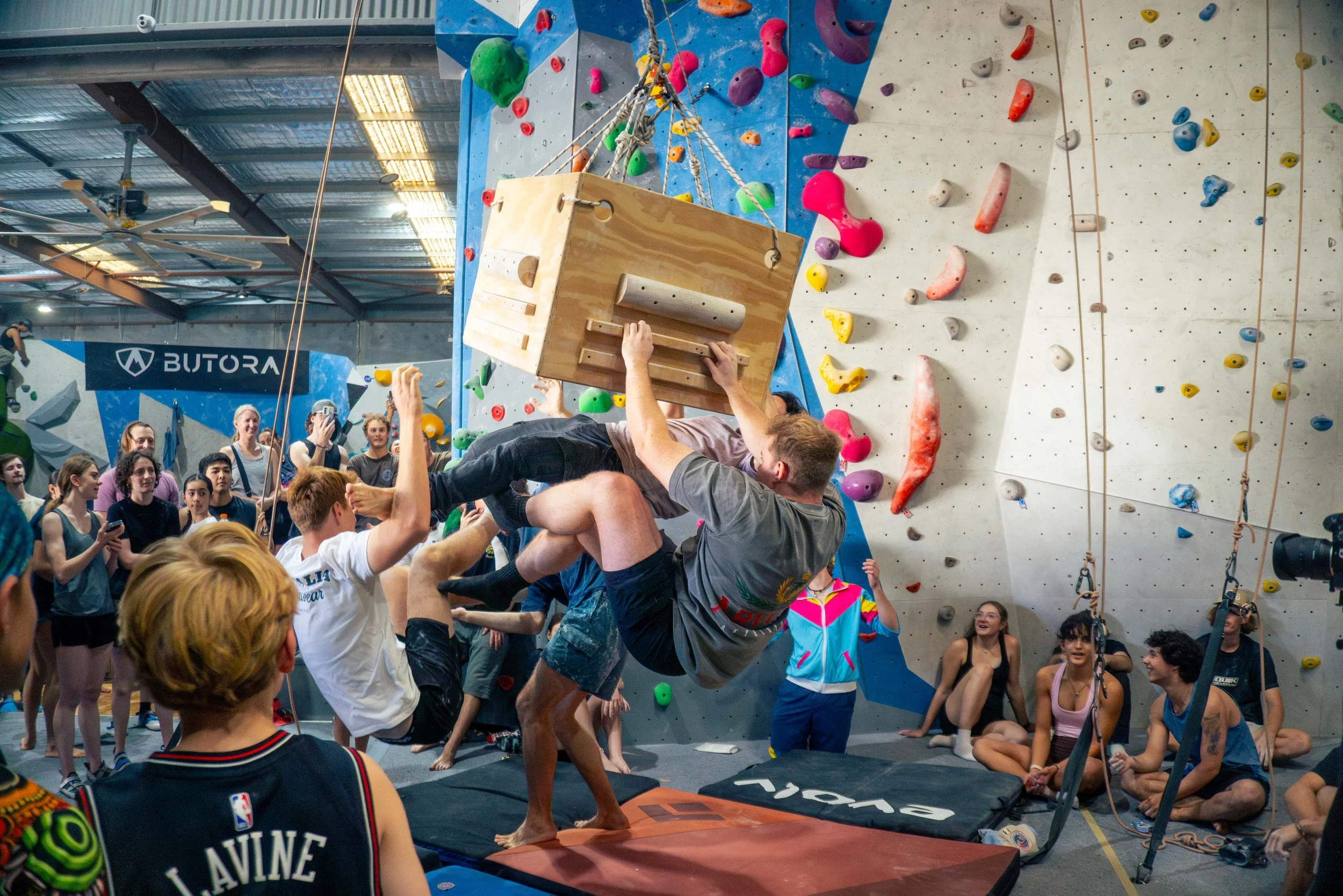 A man appears to be falling onto a padded mat while attempting to climb a wooden box at an indoor rock climbing gym. Several people are watching and smiling in the background. The climbing wall has various colorful holds.