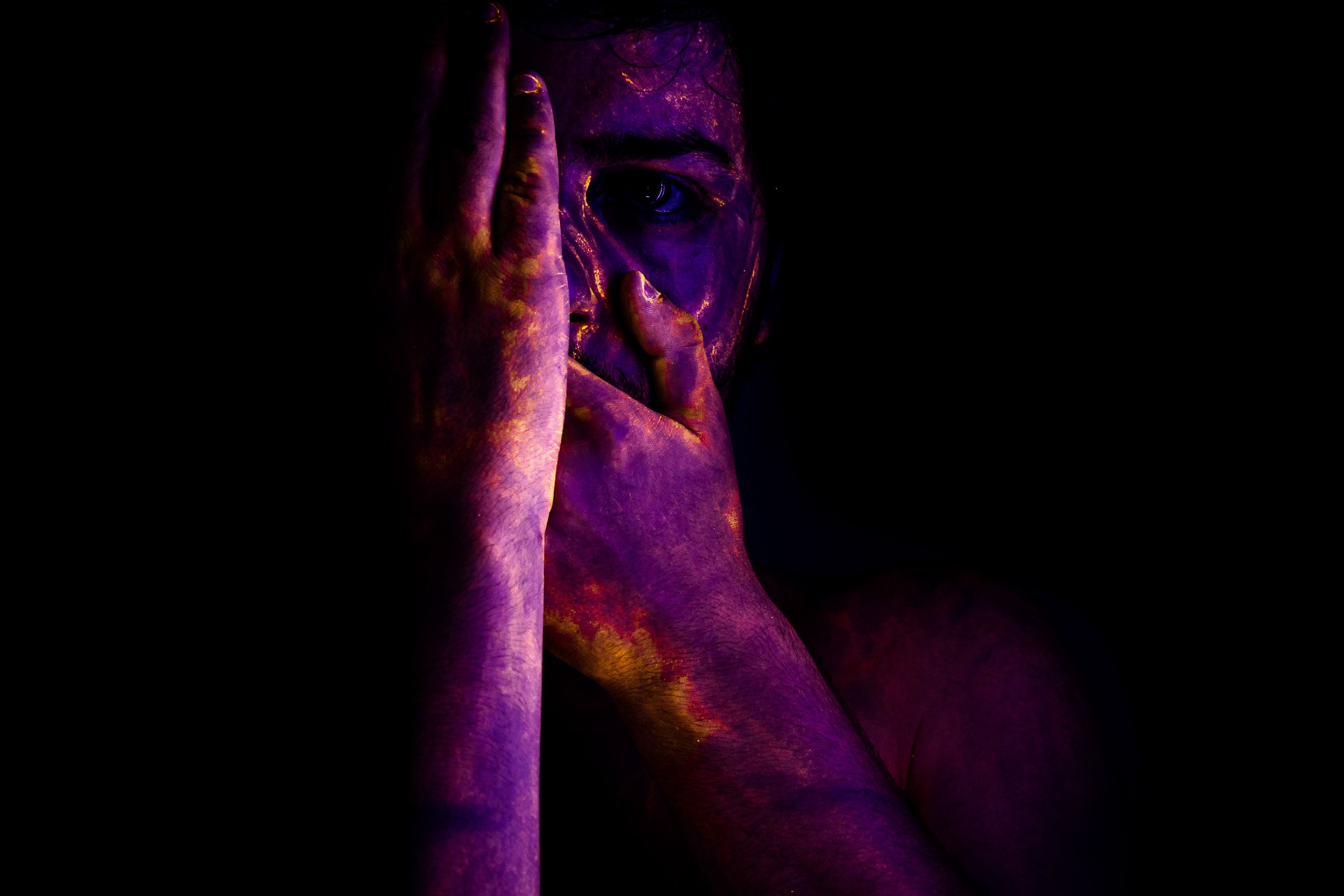 Close-up of a person's face and hand painted in vibrant purple and gold colors, with one eye visible and the person's finger held vertically near their face.
