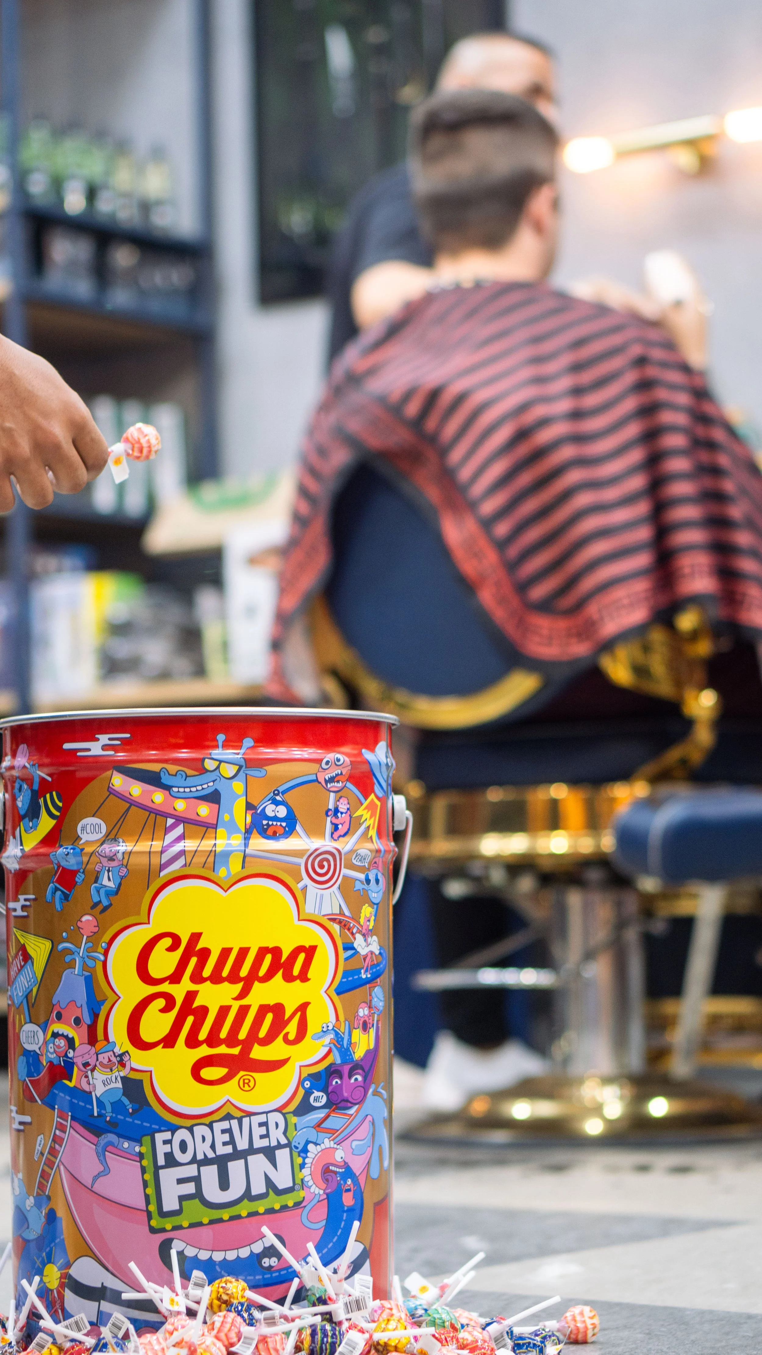 A large colorful Chupa Chups candy container labeled "Forever Fun" with various cartoon characters, surrounded by lollipops and candies on a table. In the background, two people are sitting, one with a red and black striped shirt, and another person 