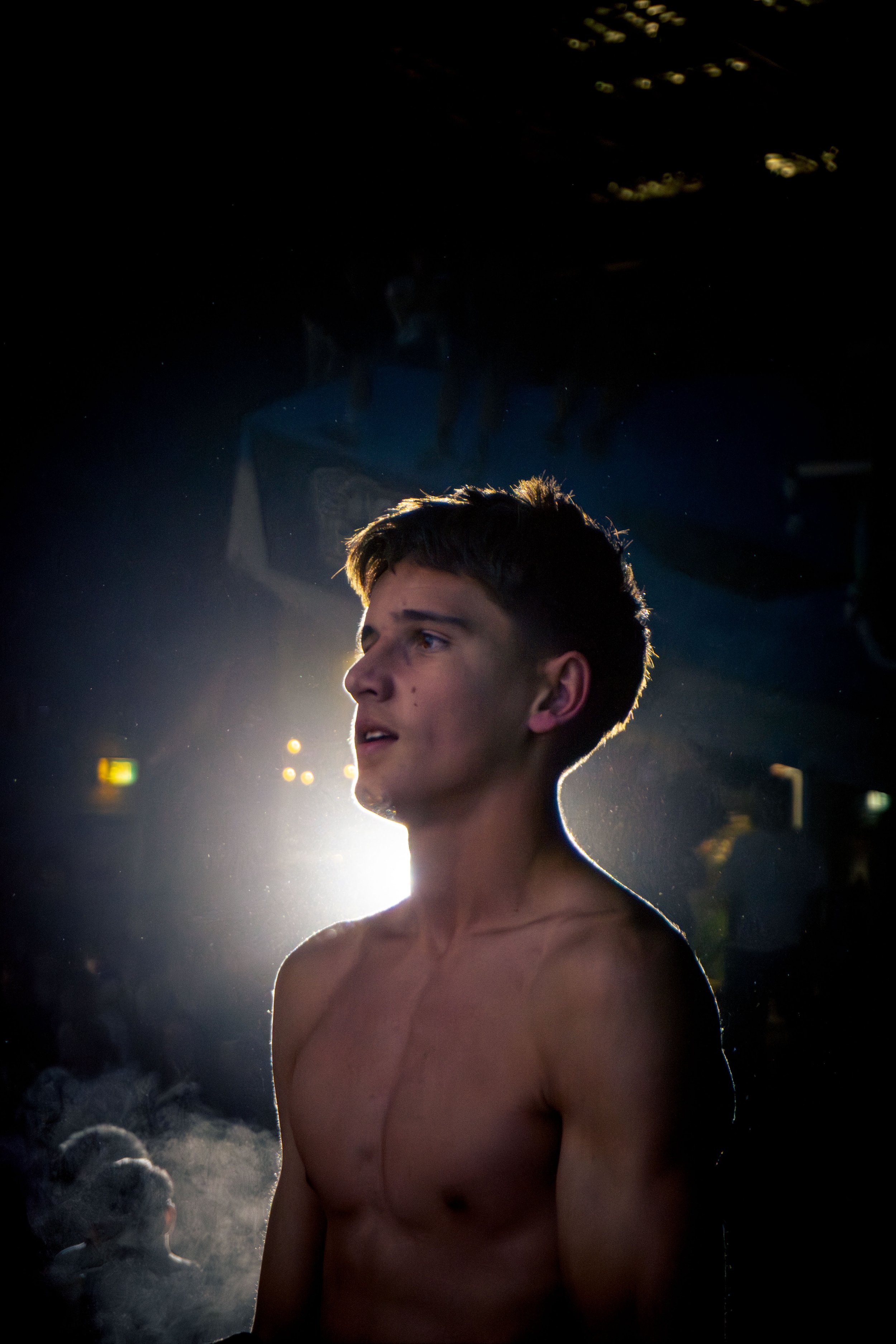 A young shirtless man with short hair standing outdoors at night, illuminated from behind by bright artificial light, with another person visible in the background.